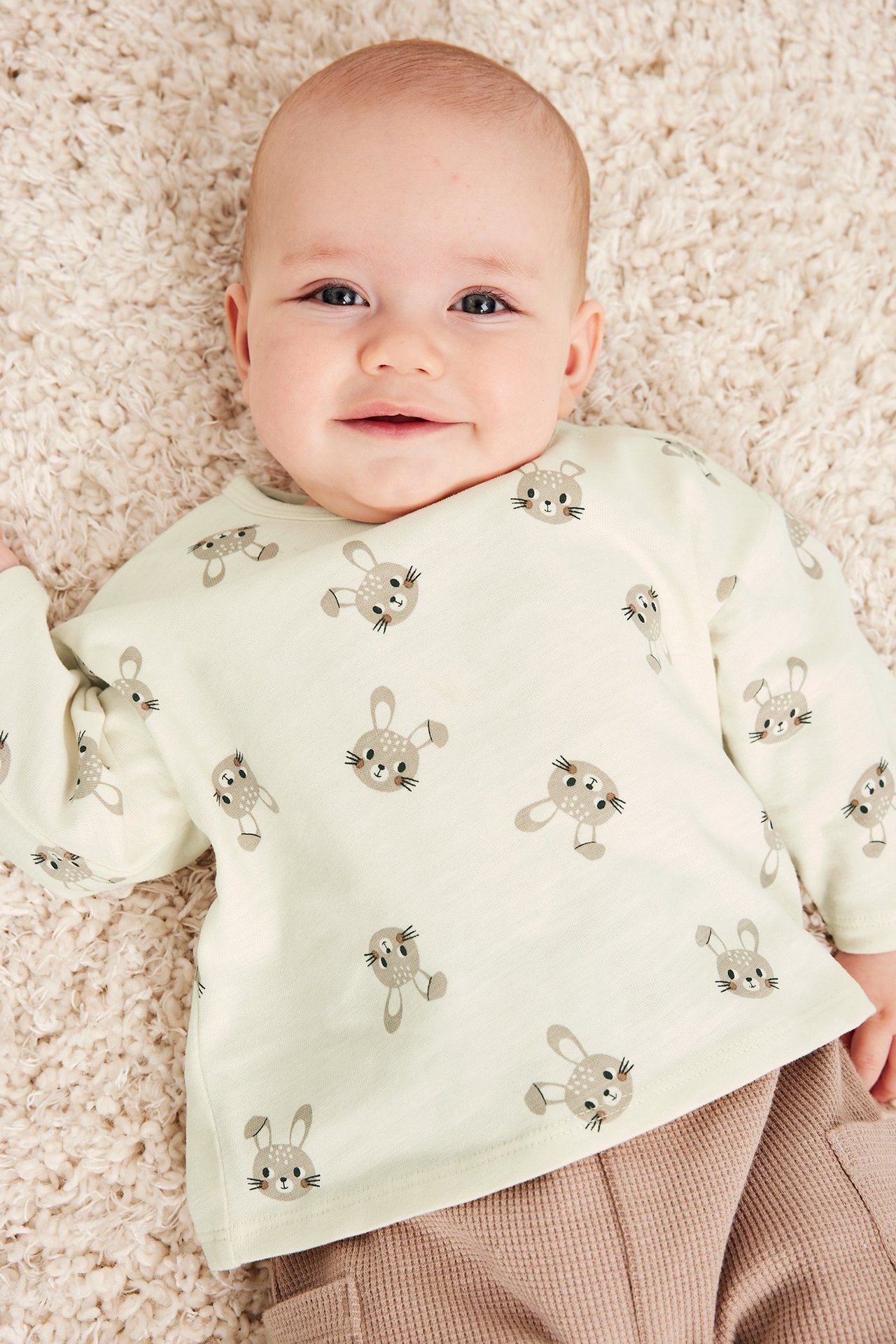 Widok z góry na niemowlę w kremowej bawełnianej koszulce z długim rękawem w króliczki i brązowych spodniach.