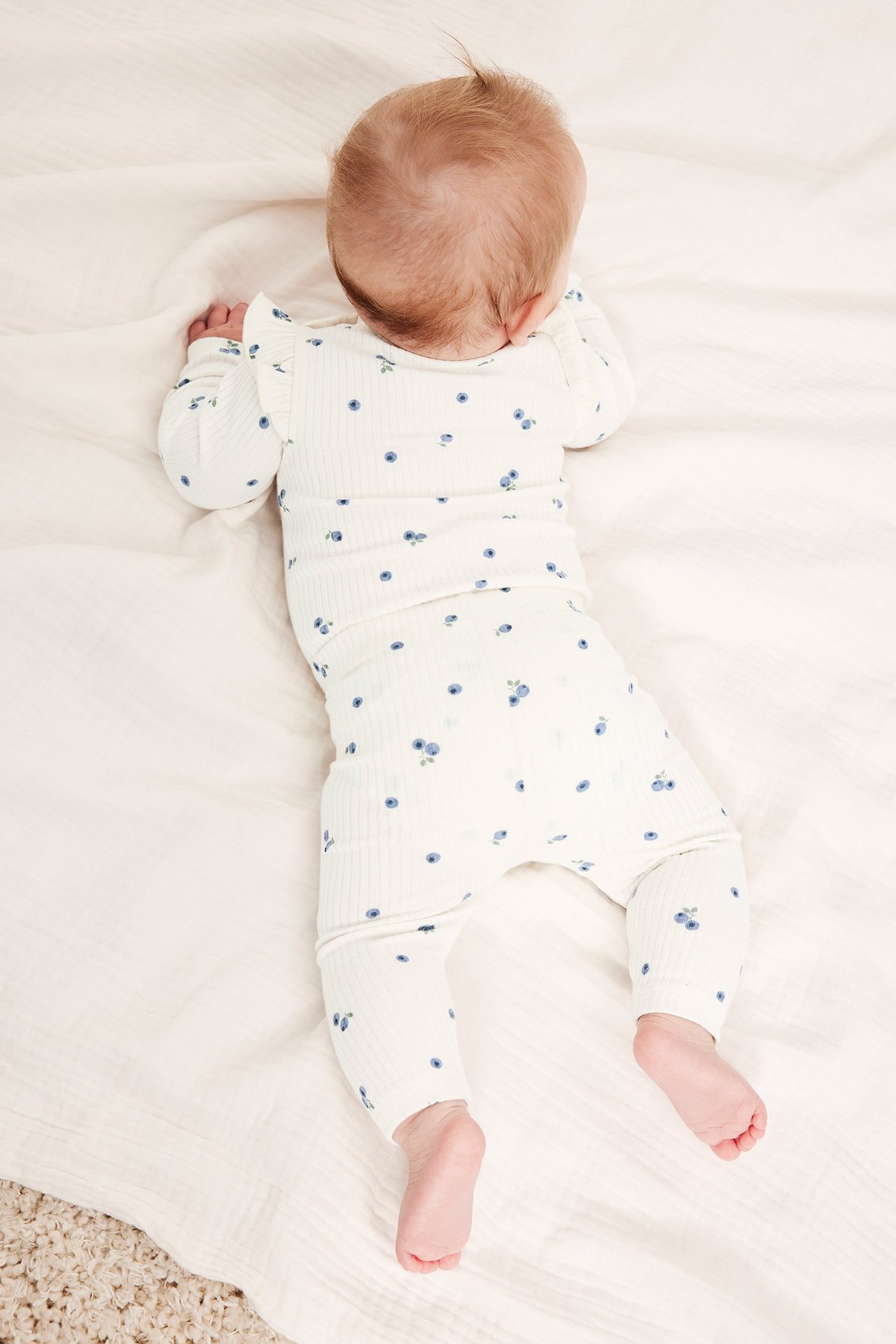 Ryggvisning av baby i offwhite ribbet body med blåbærtrykk og volanger på skuldrene.