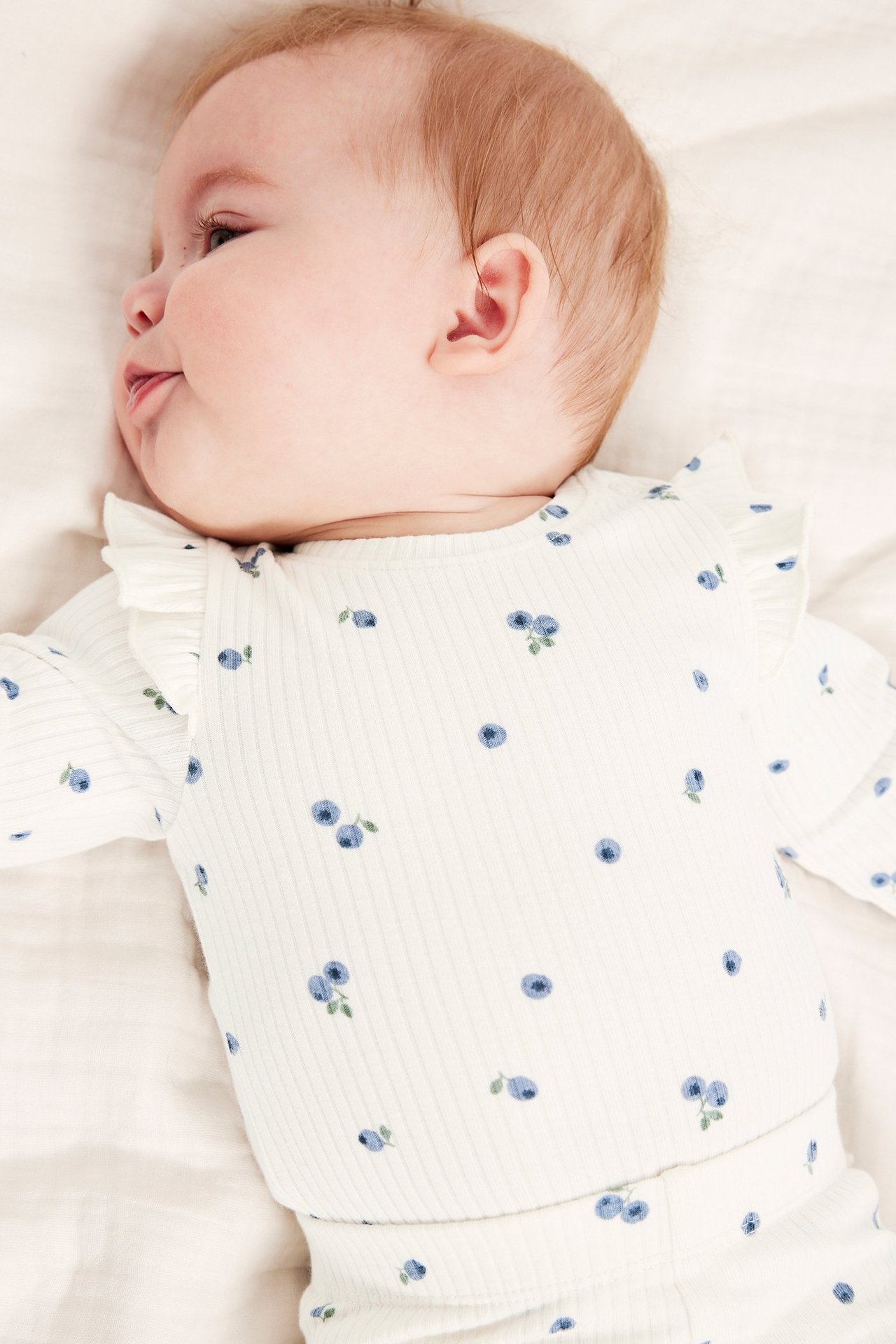Nærbilde, sidevisning av en baby i en offwhite ribbet body med blått blåbærtrykk og volangskuldre.