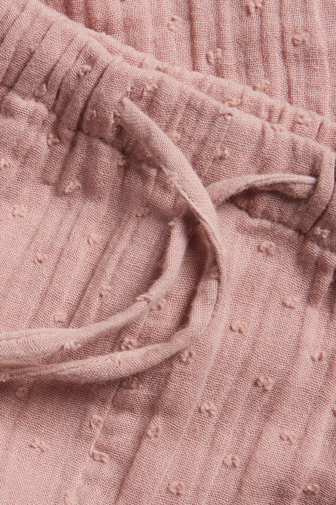 Närbild av rosa vävda bomullsbyxor med resår i midjan och dragsko, med prickig textur.