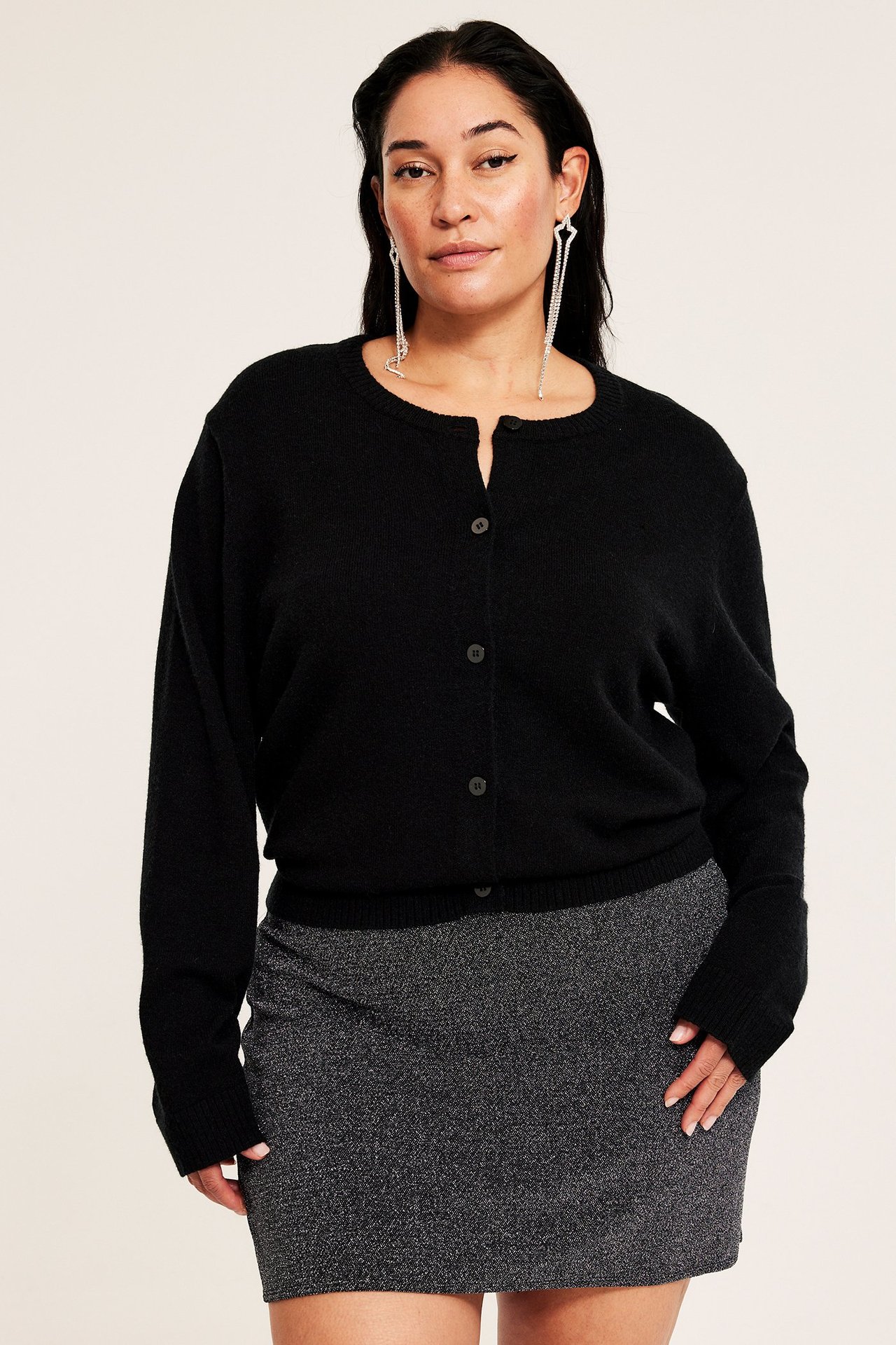 Widok z przodu kobiety w krótkiej, czarnej spódnicy z brokatowej dzianiny, czarnym kardiganie i długich kolczykach.