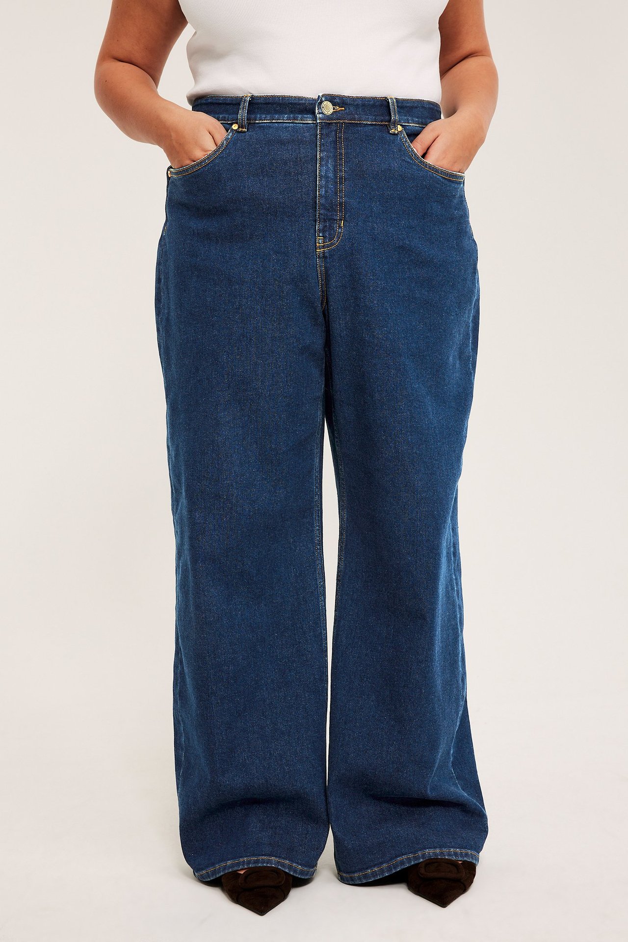 Framifrån: kvinna i vida, högmidjade blå denimjeans, händer i fickorna, med vit topp och bruna skor.