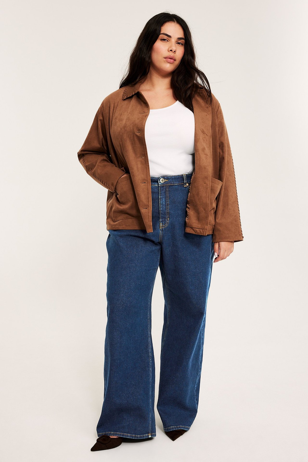 Framifrån: kvinna i blå vida raka högmidjade jeans, vit topp, brun jacka och svarta skor.