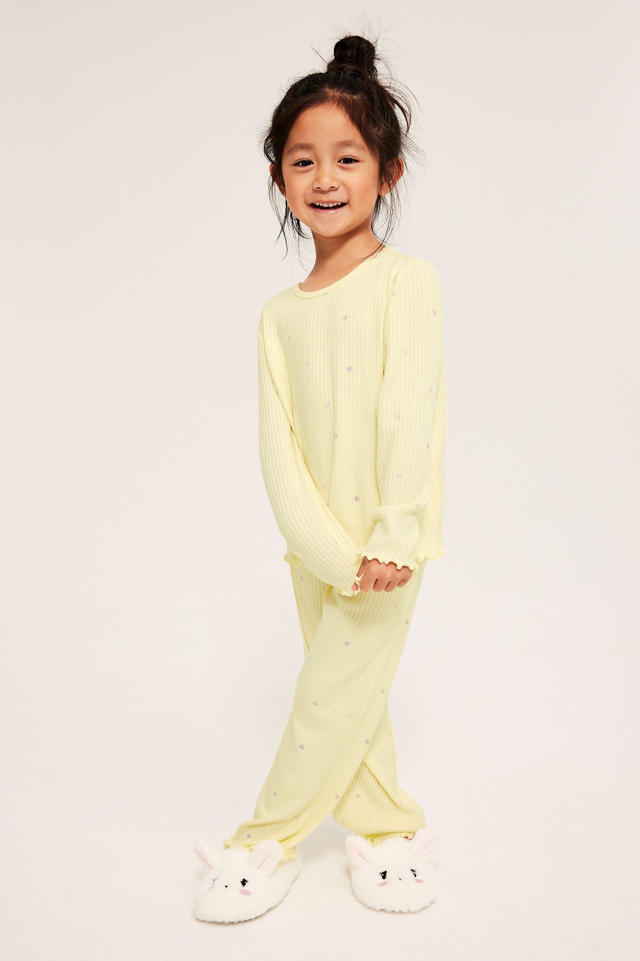 Front view av ett barn i ljusgul ribbad pyjamas med små hjärtan och volangkanter, samt vita kanintofflor.