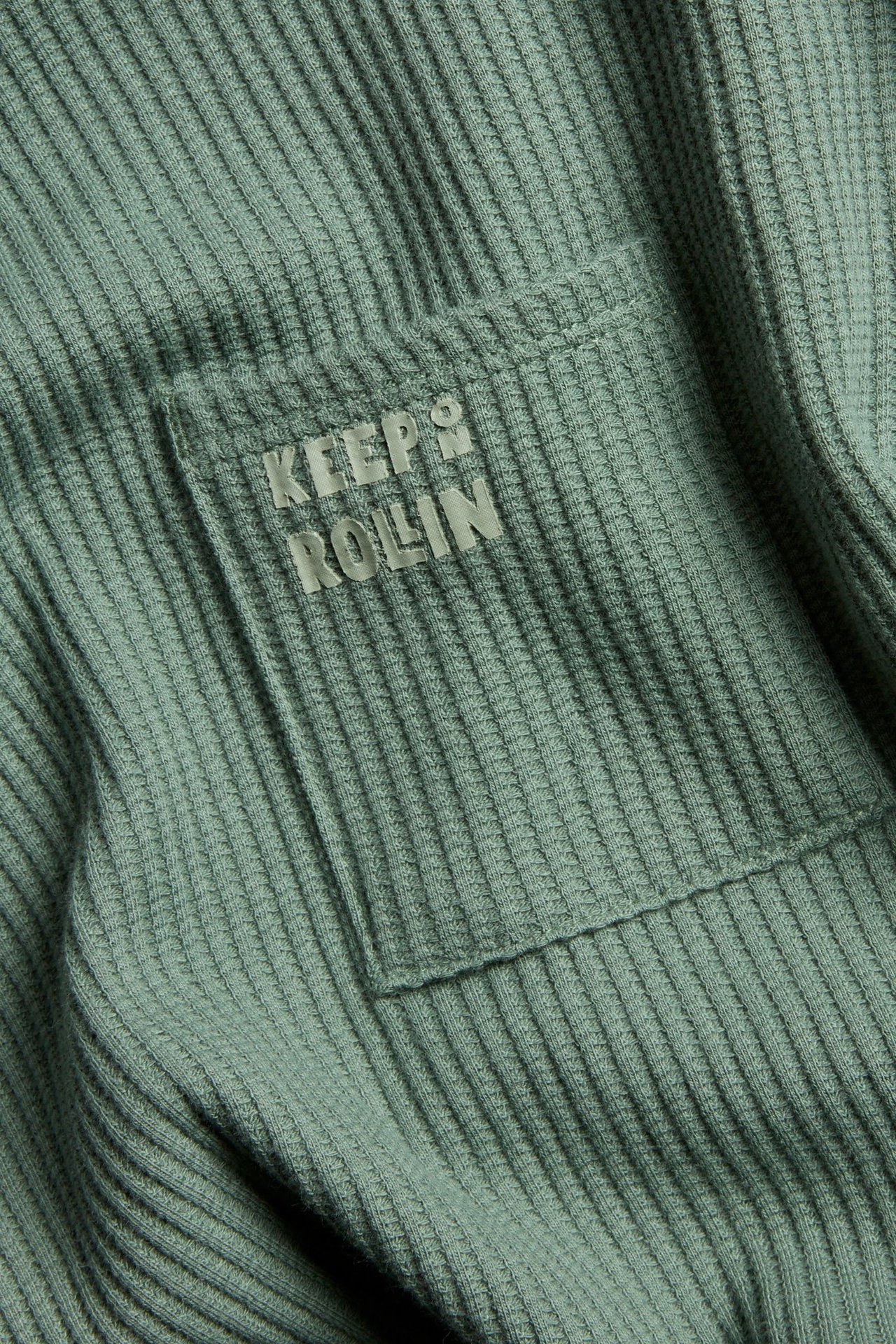 Zbliżenie na ciemnozielony, waflowy T-shirt z długim rękawem, z kieszenią na piersi i nadrukiem "KEEP ROLLIN".