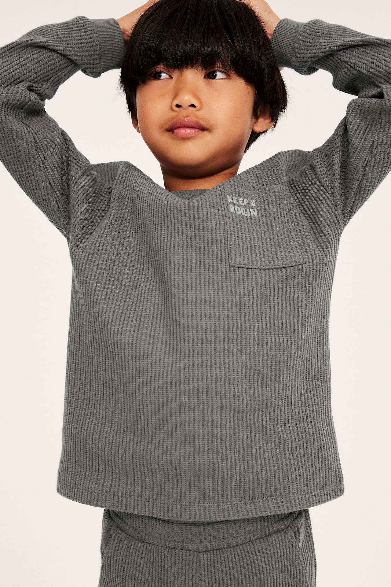 Forfra: Et barn har på seg en grå vaffelstrikket langermet t-skjorte med brystlomme og matchende bukser.
