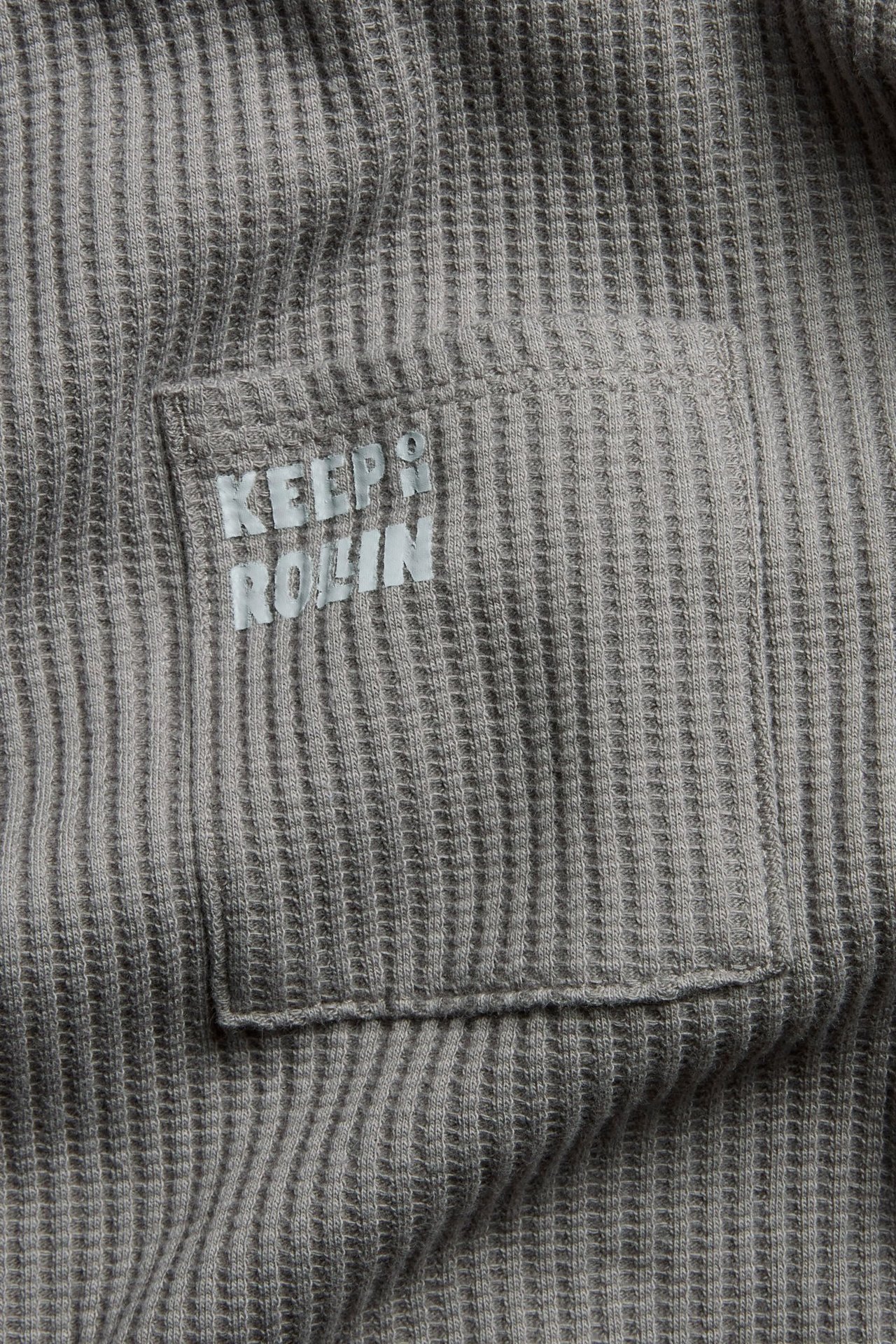 Widok z przodu: szara bawełniana koszulka z długim rękawem, waflowa struktura, kieszeń z napisem 'KEEP ROLLIN'.