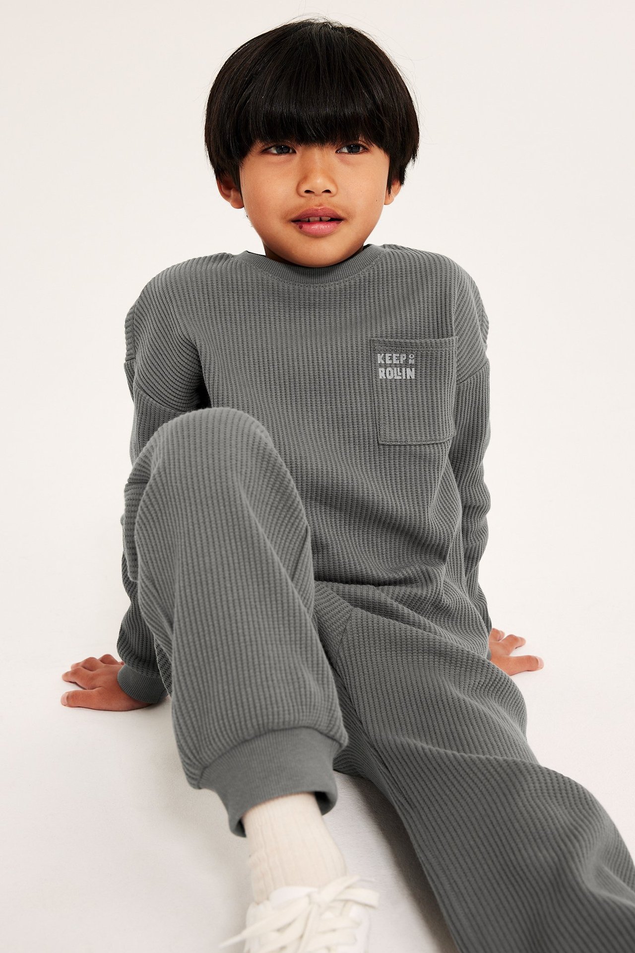 Frontbilde av et barn i en grå vaffelstrikket langermet t-skjorte med brystlomme, matchende bukser og hvite joggesko.