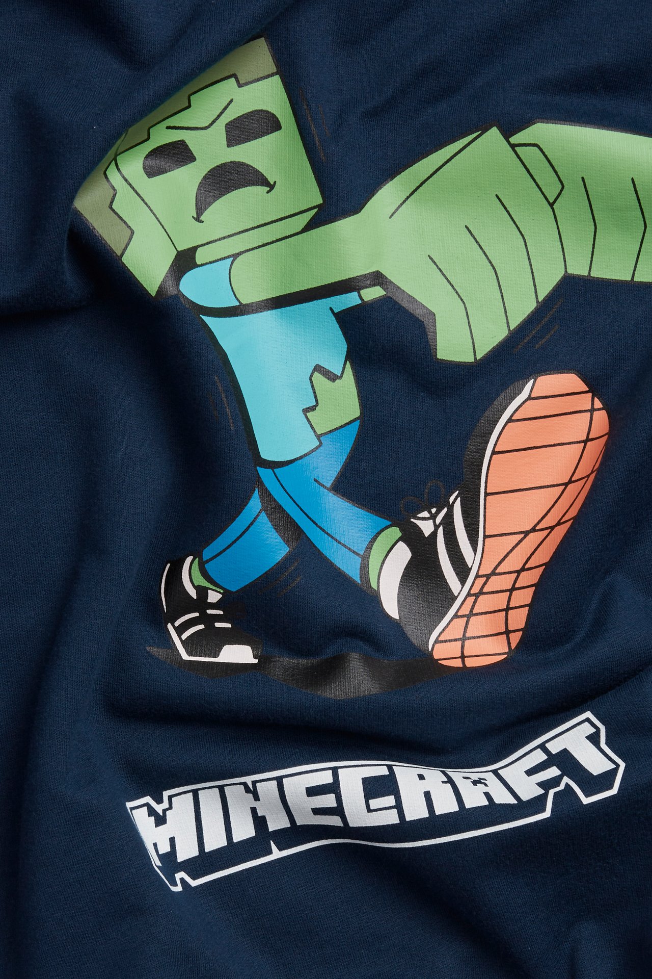Nærbilde av en mørkeblå bomullshettegenser for barn med stort grønt Minecraft zombie-trykk og hvit Minecraft-logo.
