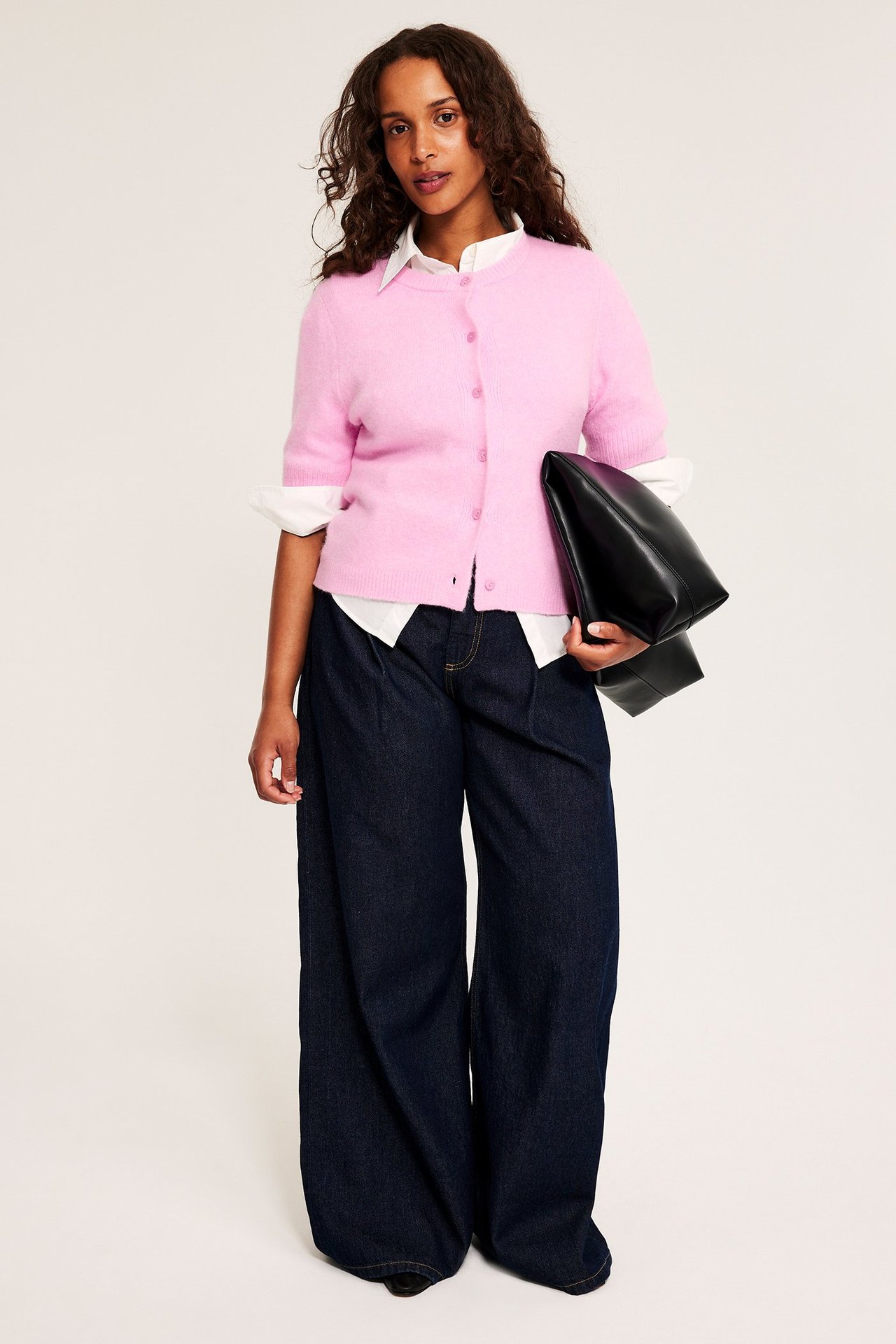 Nainen edestä: vaaleanpunainen lyhythihainen neuletakki, valkoinen paita, tummansiniset farkut, musta laukku.