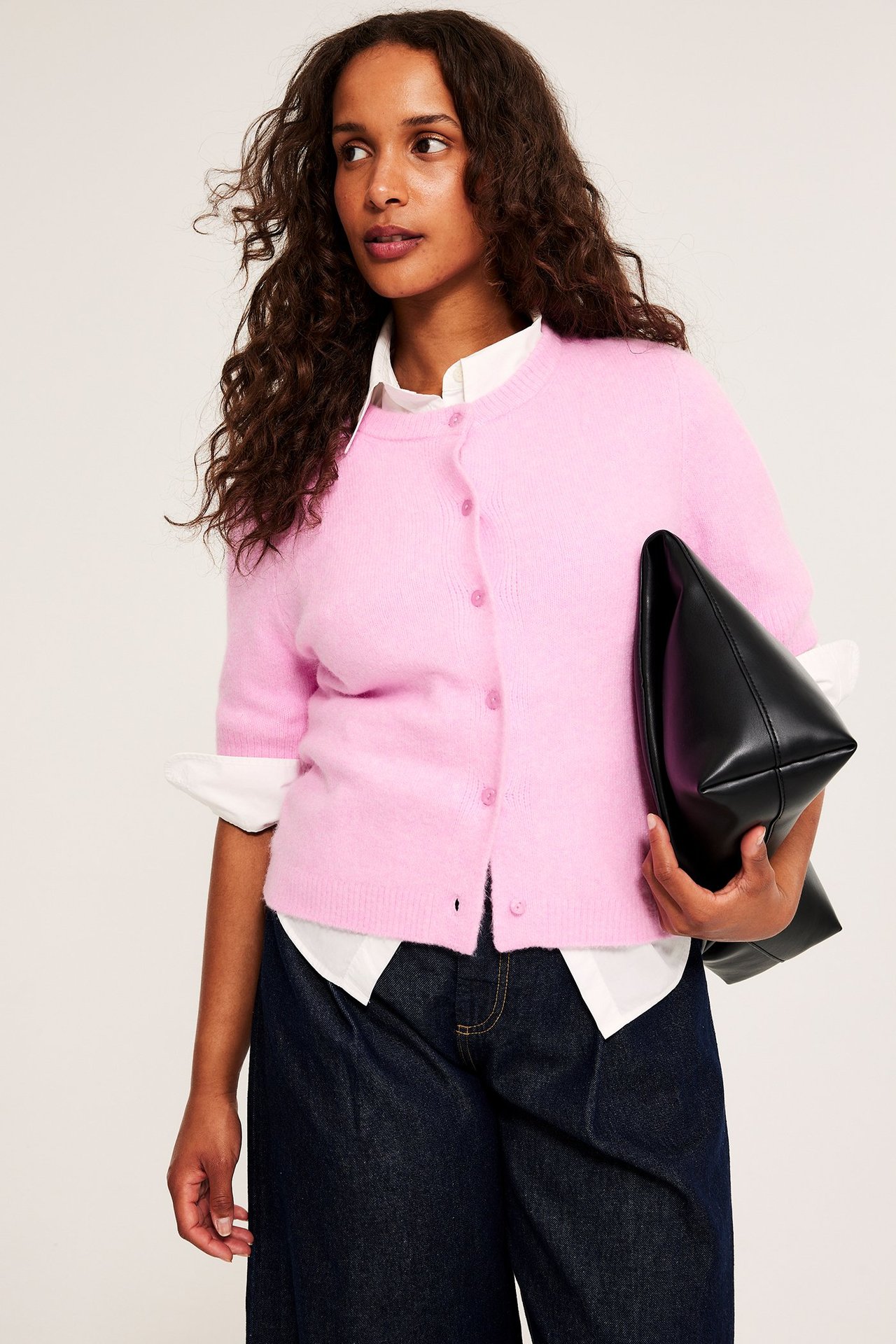Kobieta nosi różowy, krótki kardigan na białej koszuli i ciemnych dżinsach, trzymając czarną torebkę.