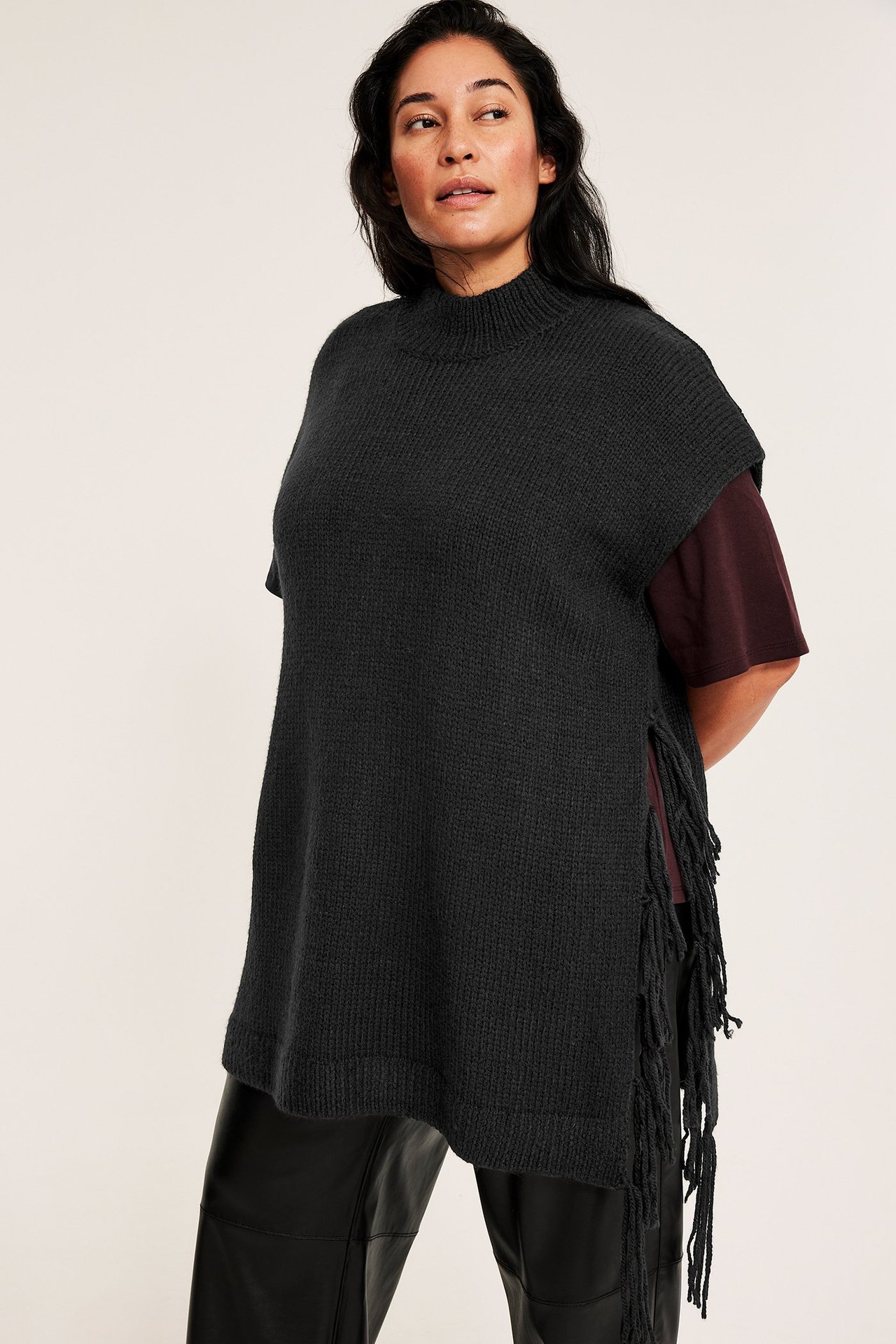 Framifrån: kvinna i mörkgrå stickad väst med fransar, över mörkröd t-shirt och svarta byxor.