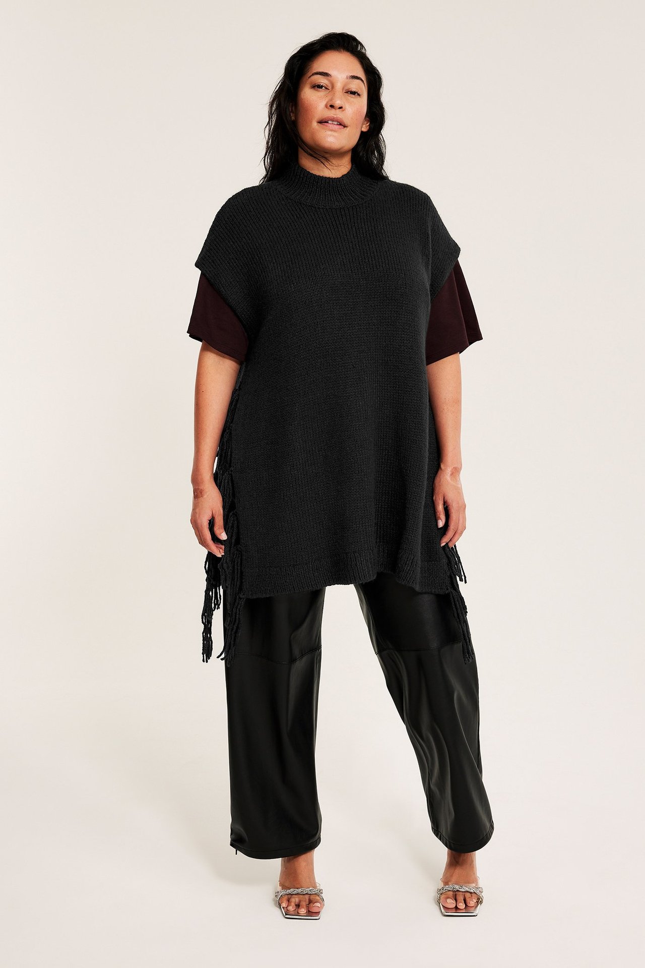 Kvinna bär en lång svart stickad väst med fransar över en mörkröd topp, svarta byxor och silversandaler.