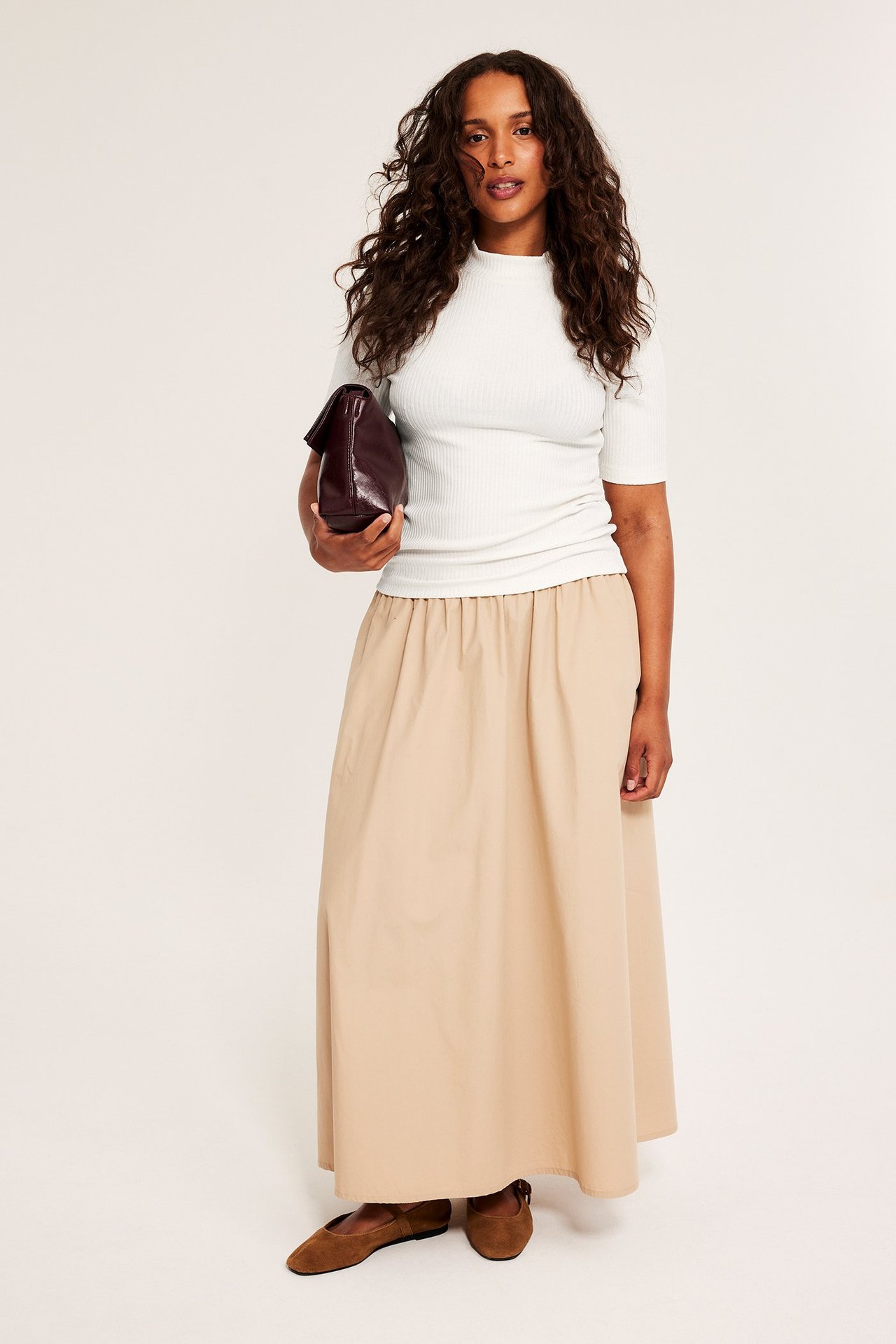 Widok z przodu: kobieta w kremowym prążkowanym topie polo, beżowej spódnicy maxi, brązowych butach, z bordową kopertówką.