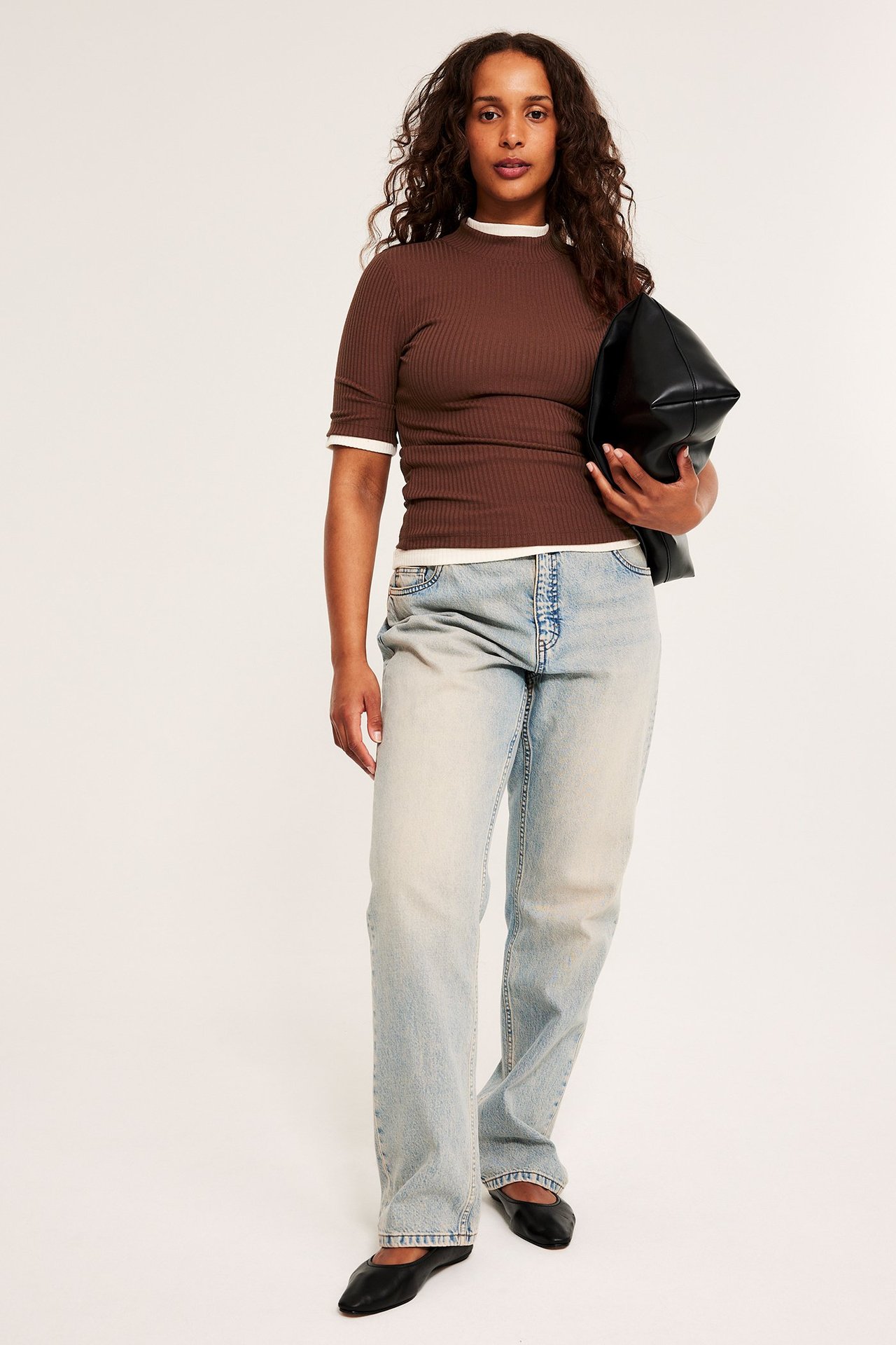 Widok z przodu: kobieta w brązowym prążkowanym topie polo, jasnych dżinsach, czarnych balerinach, trzyma czarną torebkę.