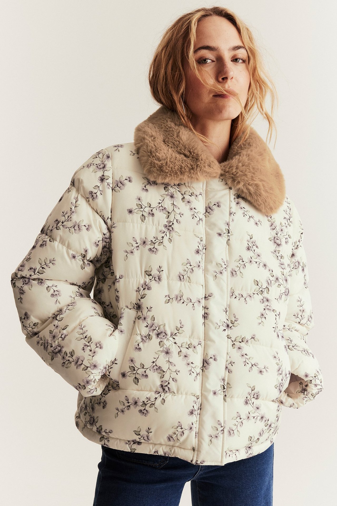 Framifrån, kvinna i ljusbeige blommig vadderad jacka med brun fuskpälskrage och mörkblå jeans.