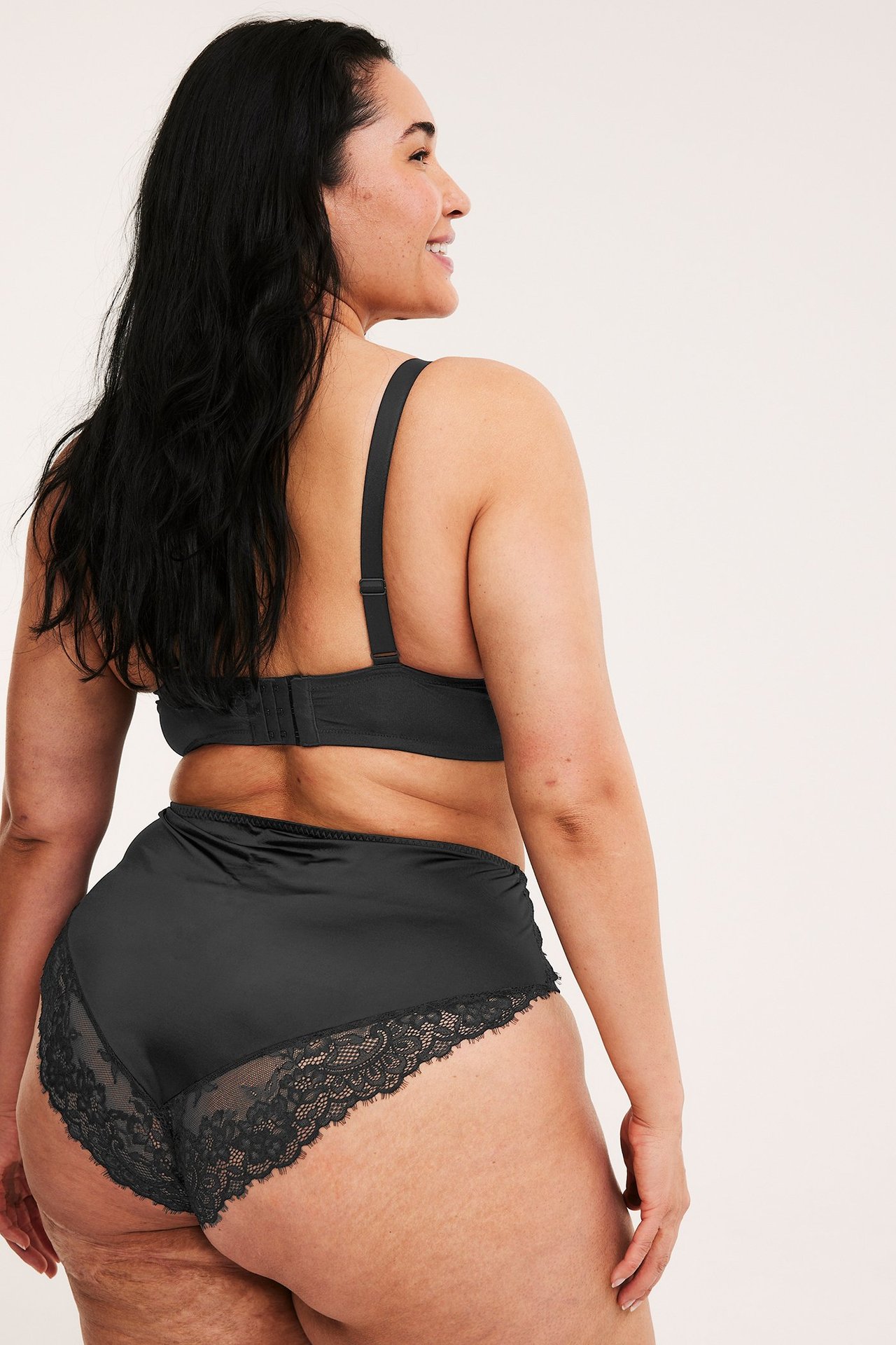 Naisen taka-sivukuva mustissa pitsireunaisissa satiinisissa cheekyalushousuissa ja rintaliiveissä.