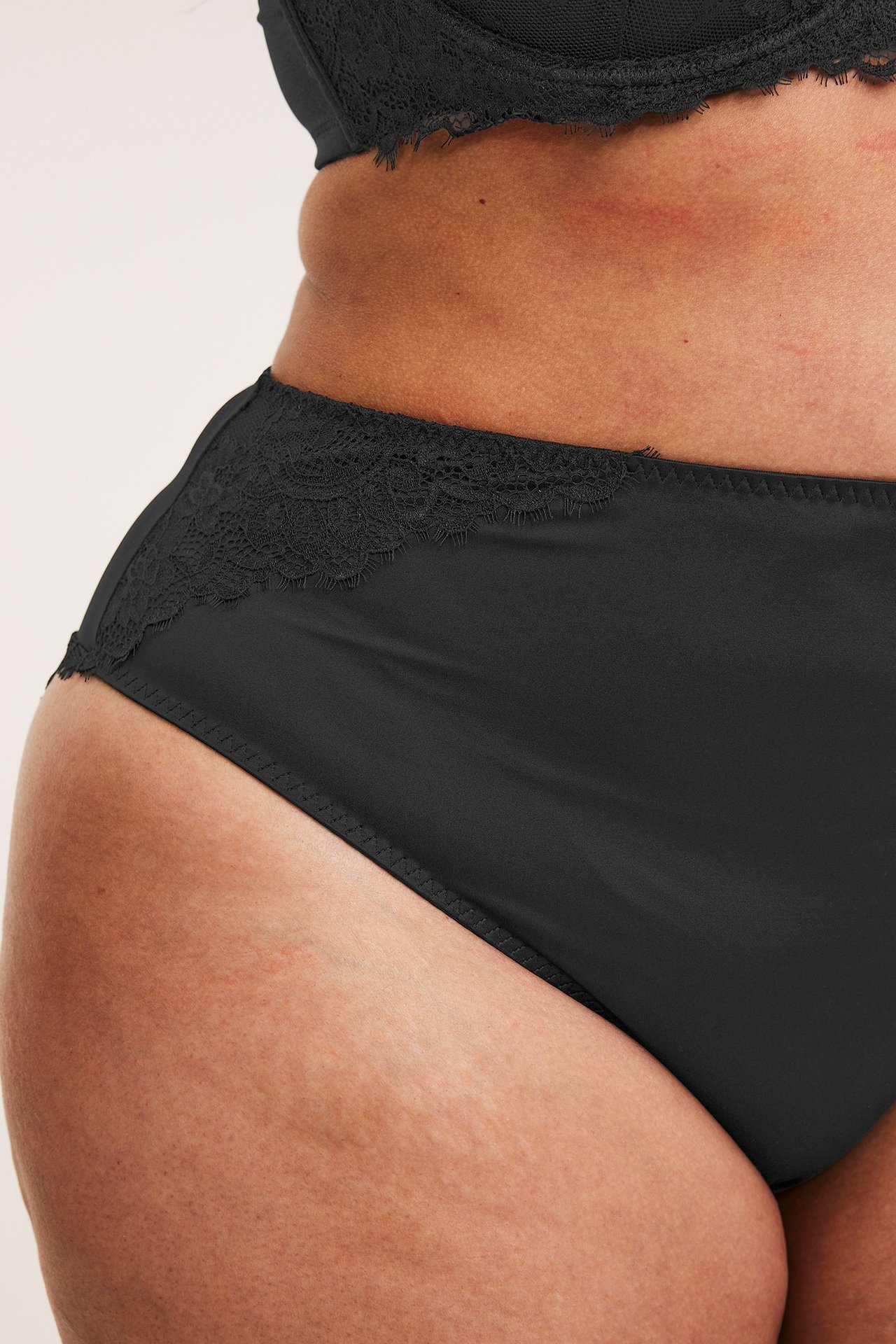 Lähikuva sivulta naisen mustat satiinialushousut pitsireunalla ja yhteensopivat rintaliivit.