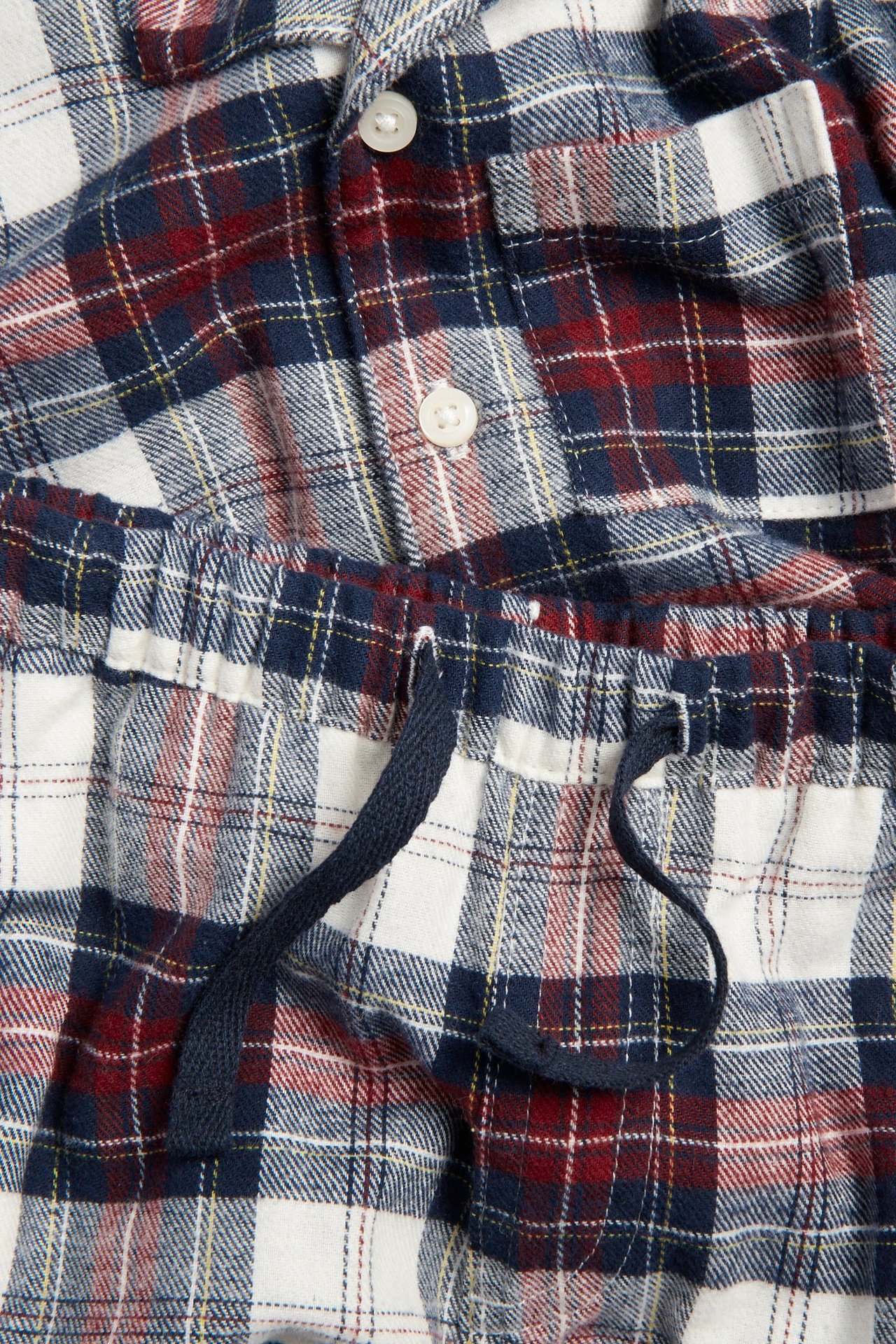 Lähikuva luonnonvalkoisesta, punaisesta ja sinisestä ruutufanellipyjamasta; napillinen yläosa taskulla, alaosassa kiristysnyöri.