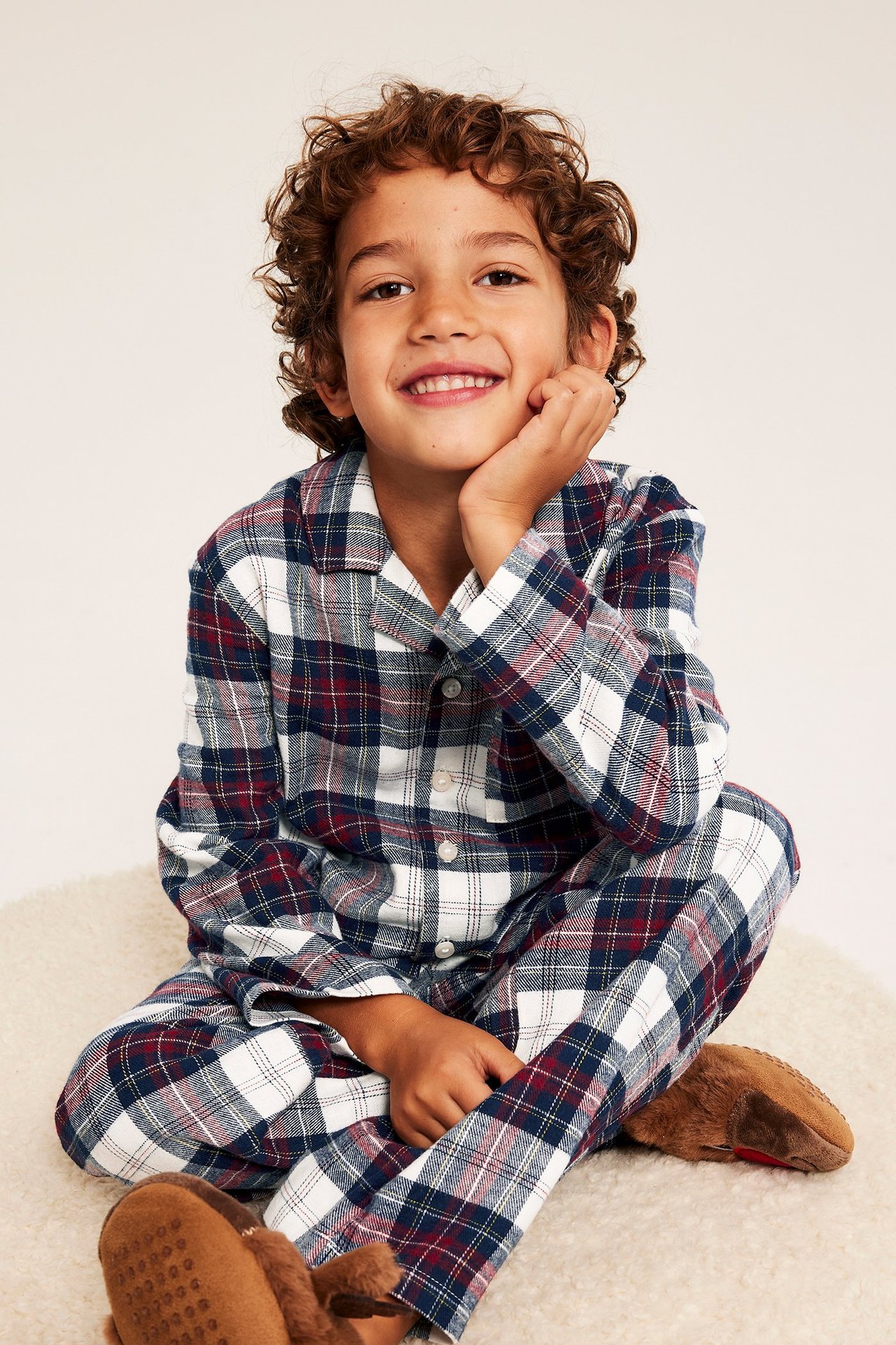 Frontvy av ett leende barn i rutig flanellpyjamas i offwhite, marinblått, rött och bruna tofflor.