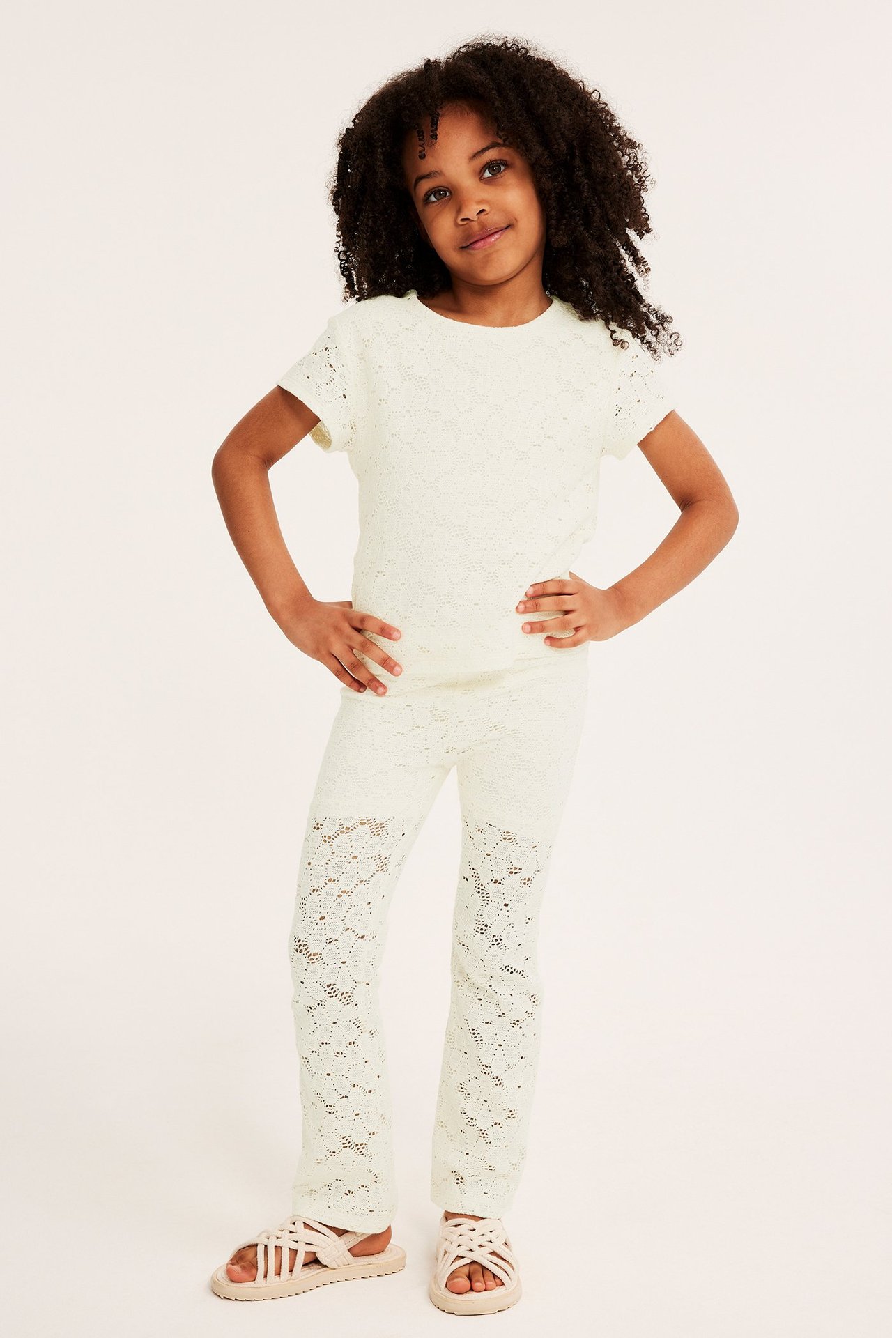 Widok z przodu dziecka w kremowych koronkowych spodniach dzwonach, pasującym topie i jasnoróżowych sandałach.