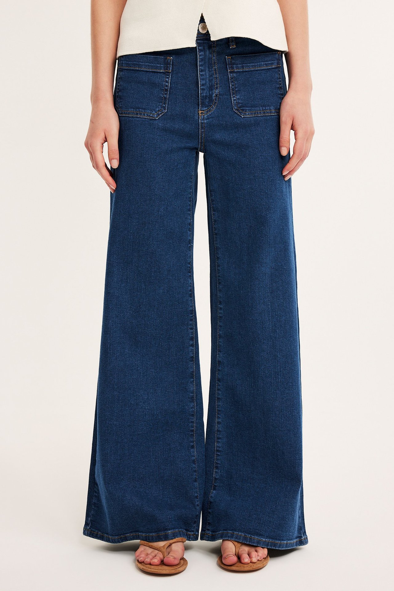 Framifrån: Kvinna i mörkblå höga vida jeans med påsydda fickor fram, ljus topp och bruna flip-flops.