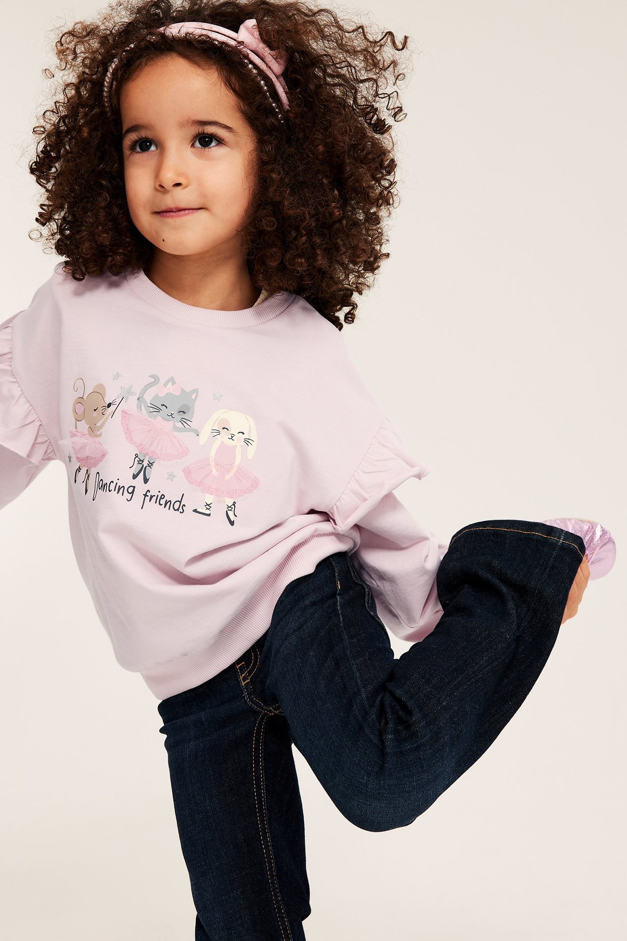 Framsida: Barn i ljusrosa volangtopp med tryck, mörkblå jeans och rosa hårband.