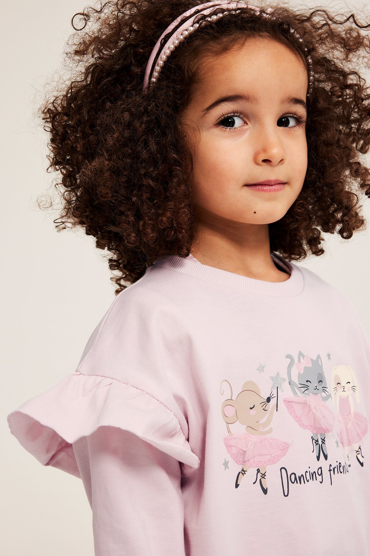 Dziecko nosi jasnoróżowy top z długim rękawem, nadrukiem zwierzęcym, falbankami i różową opaską z pereł.