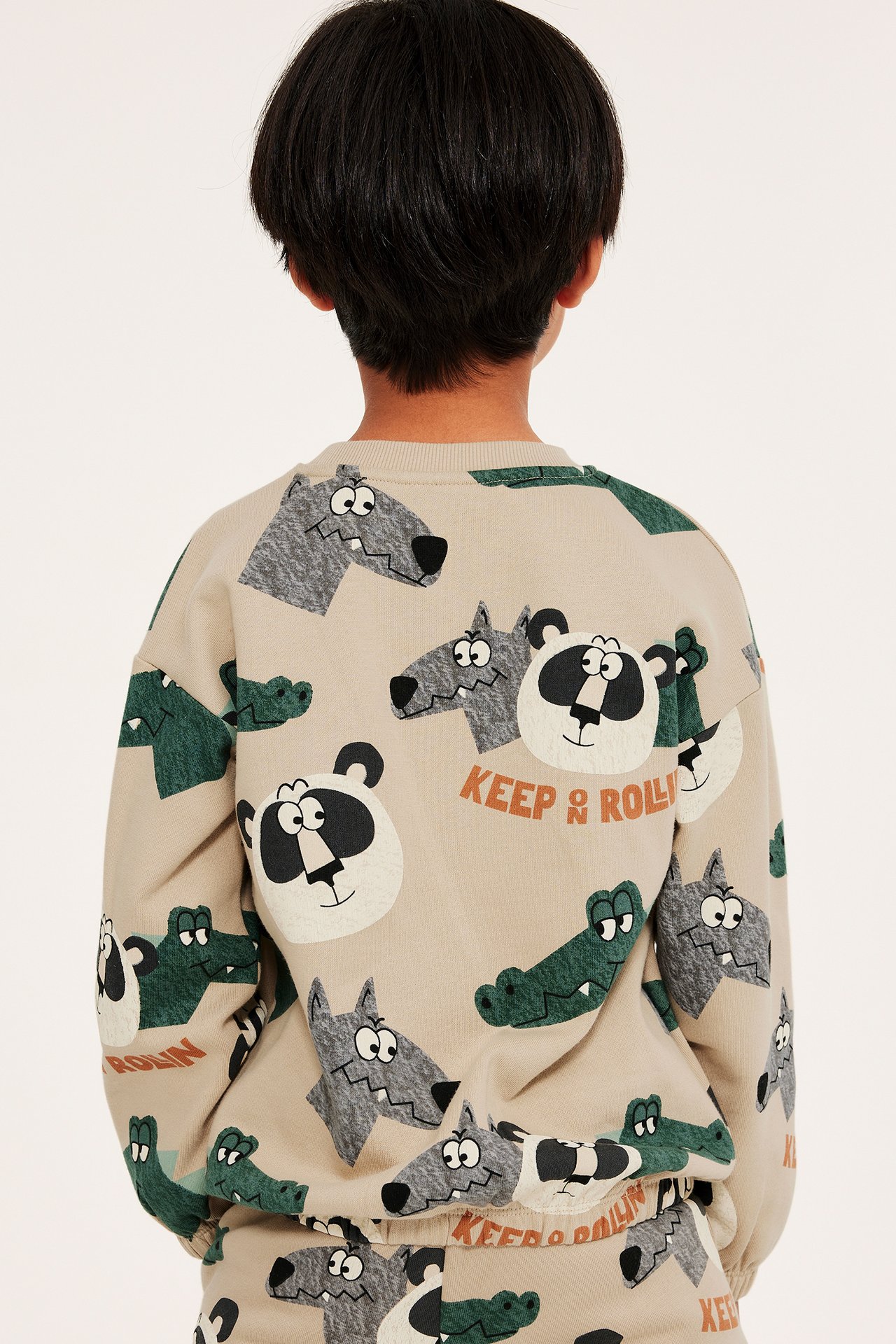 Bakifrån på ett barn i en beige bomullssweatshirt med heltäckande djurmönster och texten 'KEEP & ROLLIN''.