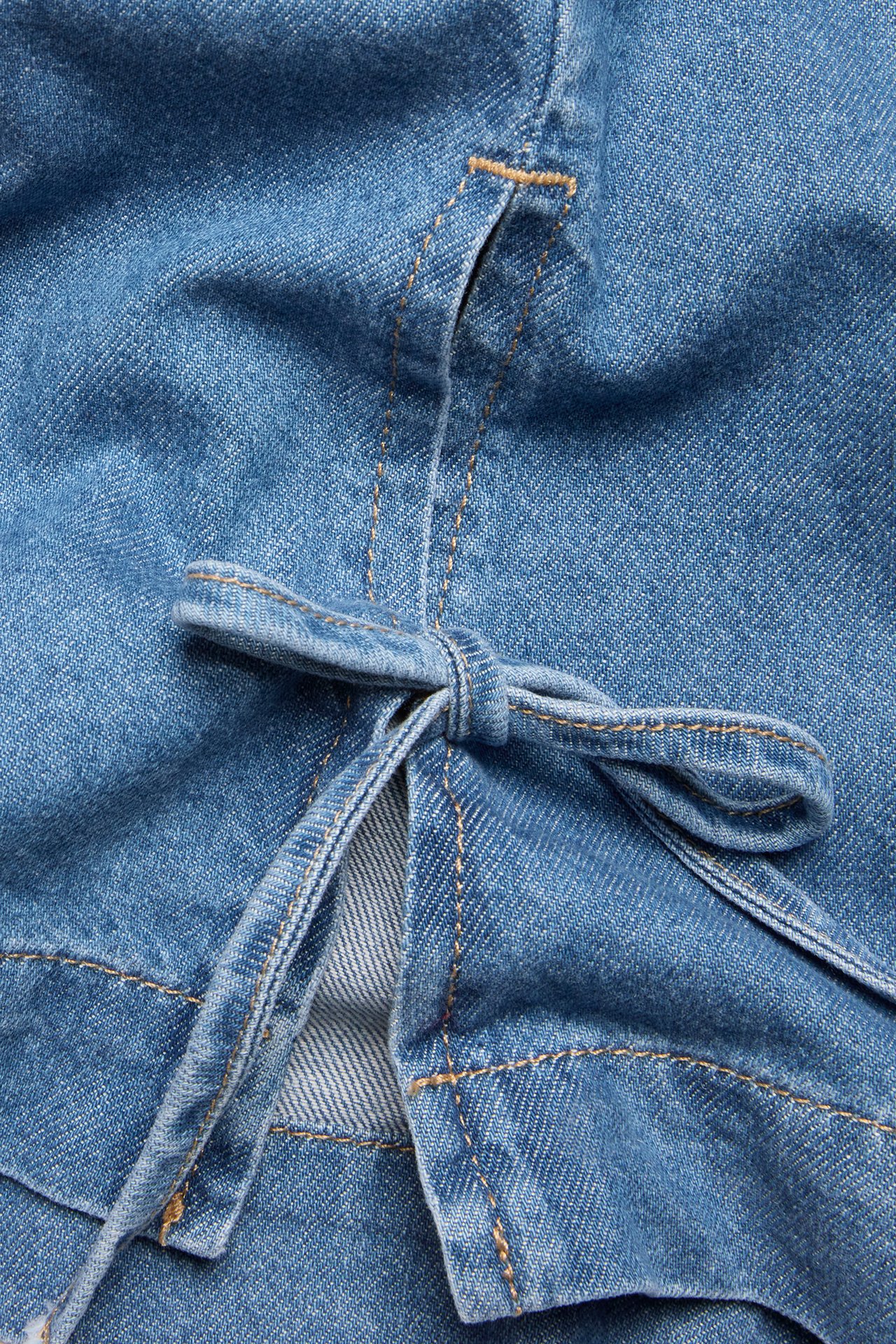 Närbild av ett ljusblått denimlinne med sidoslits och knytband, med synliga sömmar.