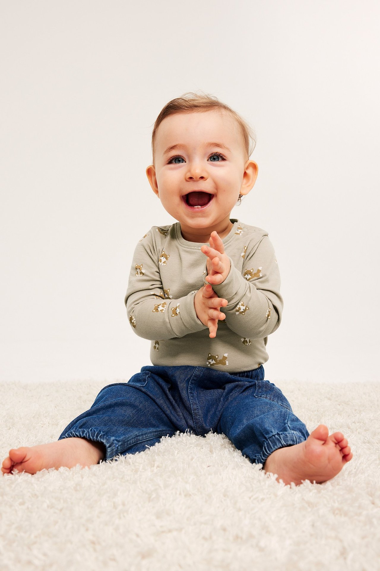 Edestä, iloinen vauva yllään vaaleanvihreä kuviollinen pitkähihainen body ja tummansiniset farkut.