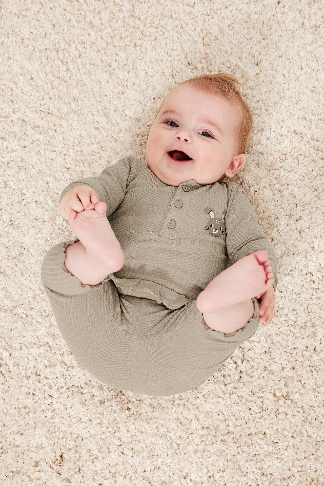 Ovenfra: glad baby i beige ribbet body med knapper foran, kanintrykk og matchende bukser.