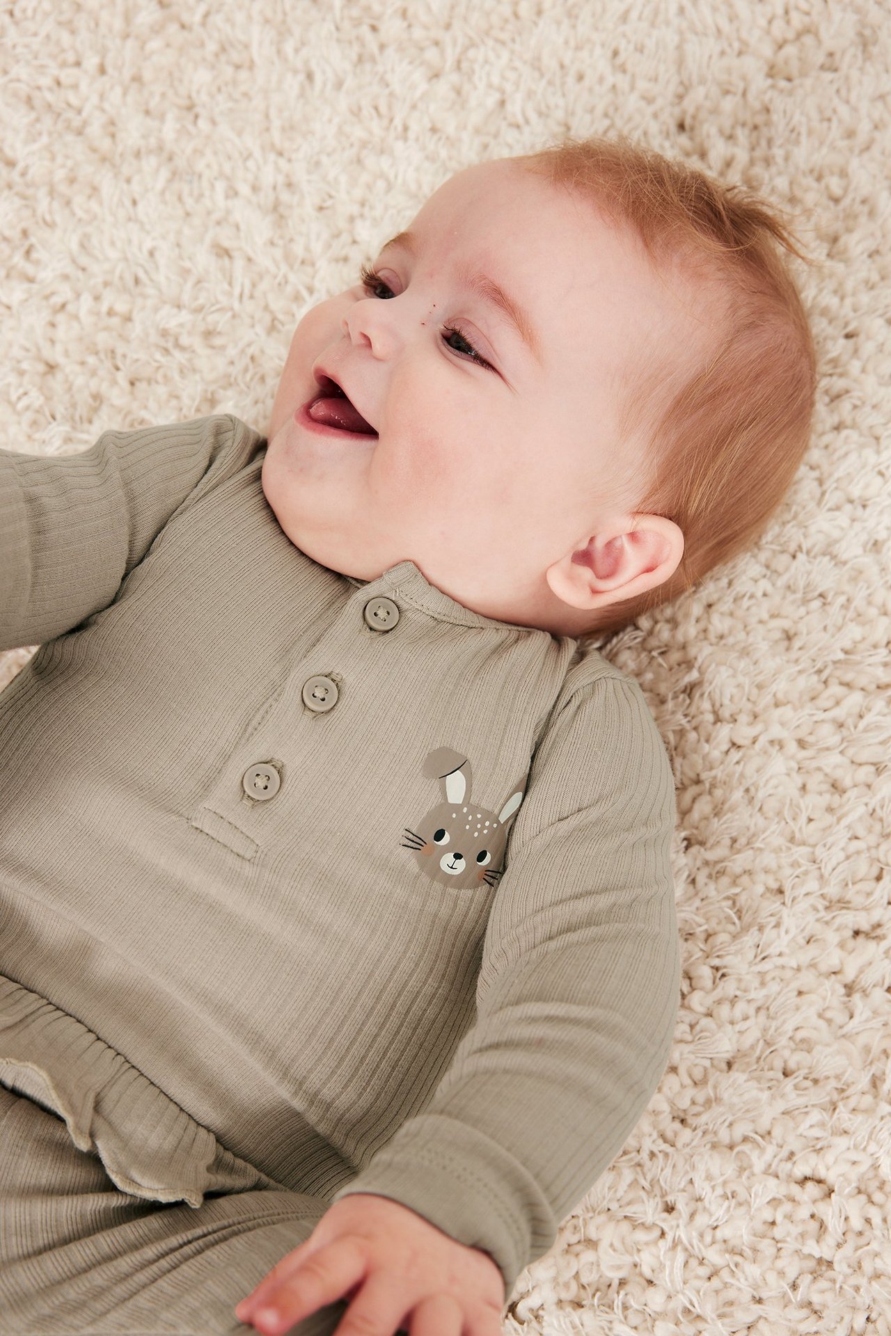 Nærbilde av smilende baby i beige ribbet body med knapper foran og kanintrykk.