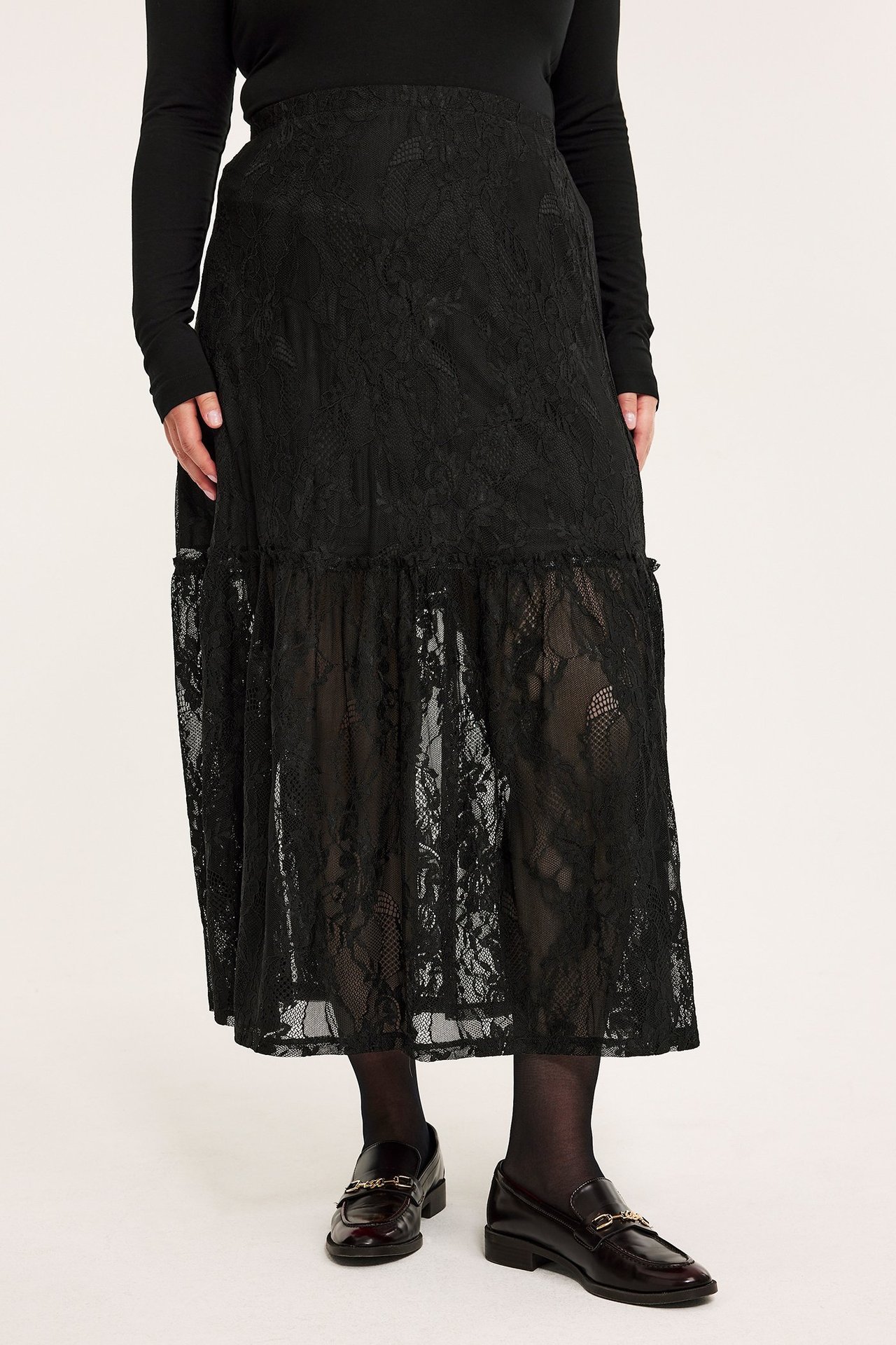Frontvy: kvinna i lång svart spetskjol med volang, svart topp och mörkbruna loafers.