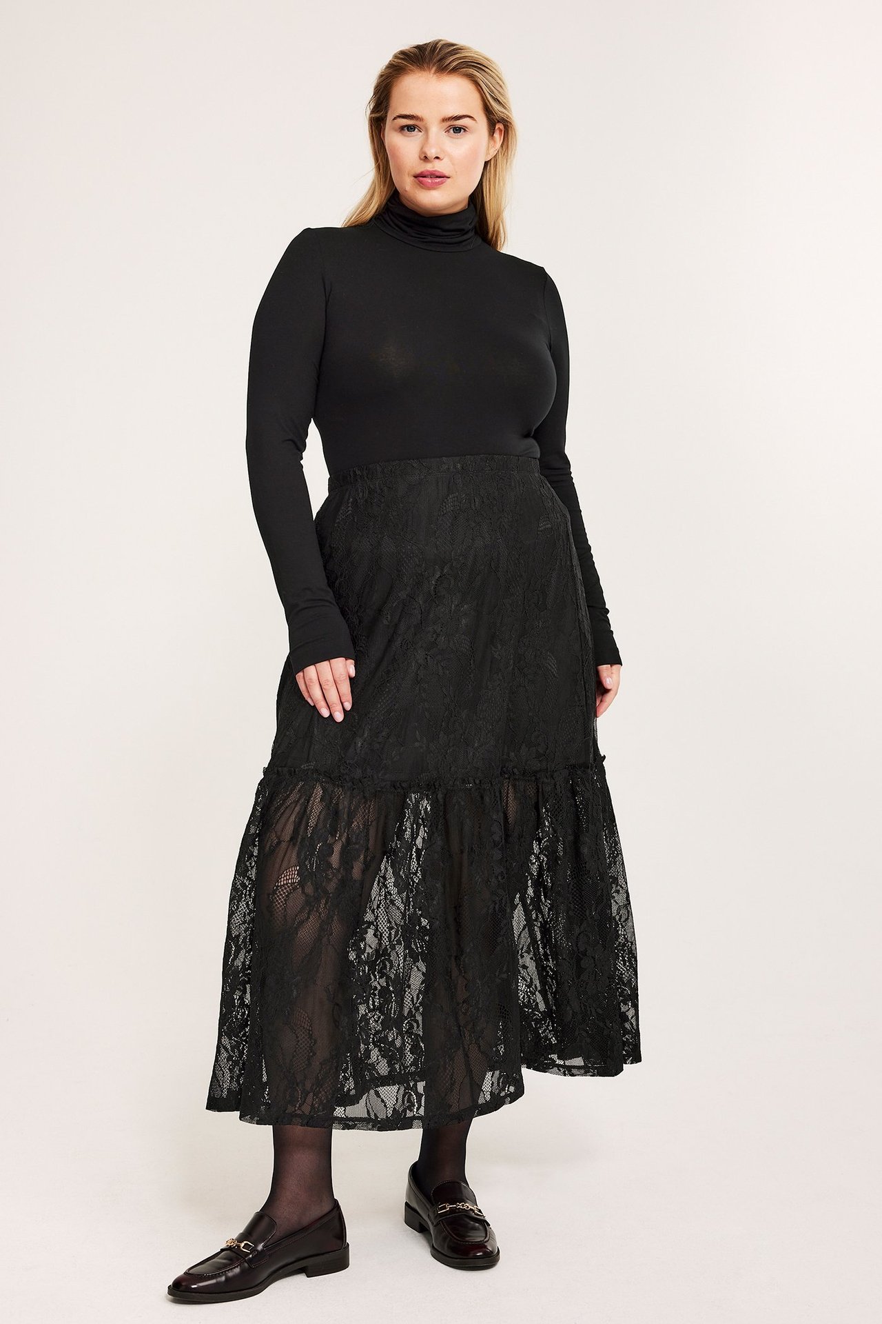 Widok z przodu: kobieta w czarnej koronkowej spódnicy midi z falbaną, czarnym golfie, rajstopach i mokasynach.