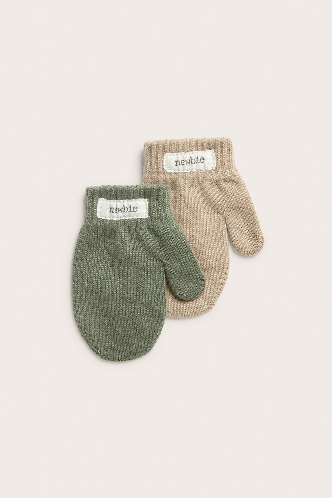 Ylhäältä näkymä kahdesta neulotusta vauvan lapasesta, toinen tummanvihreä ja toinen beige, etiketeillä.