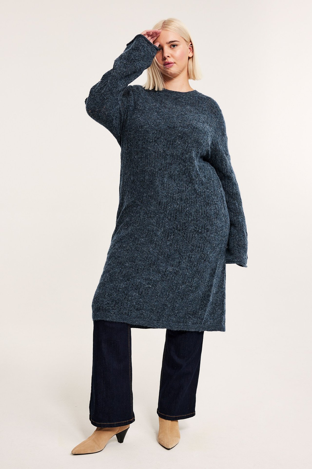 Frontvy av en kvinna i en mörkblå lös stickad klänning över mörkblå byxor och beiga ankelstövlar.