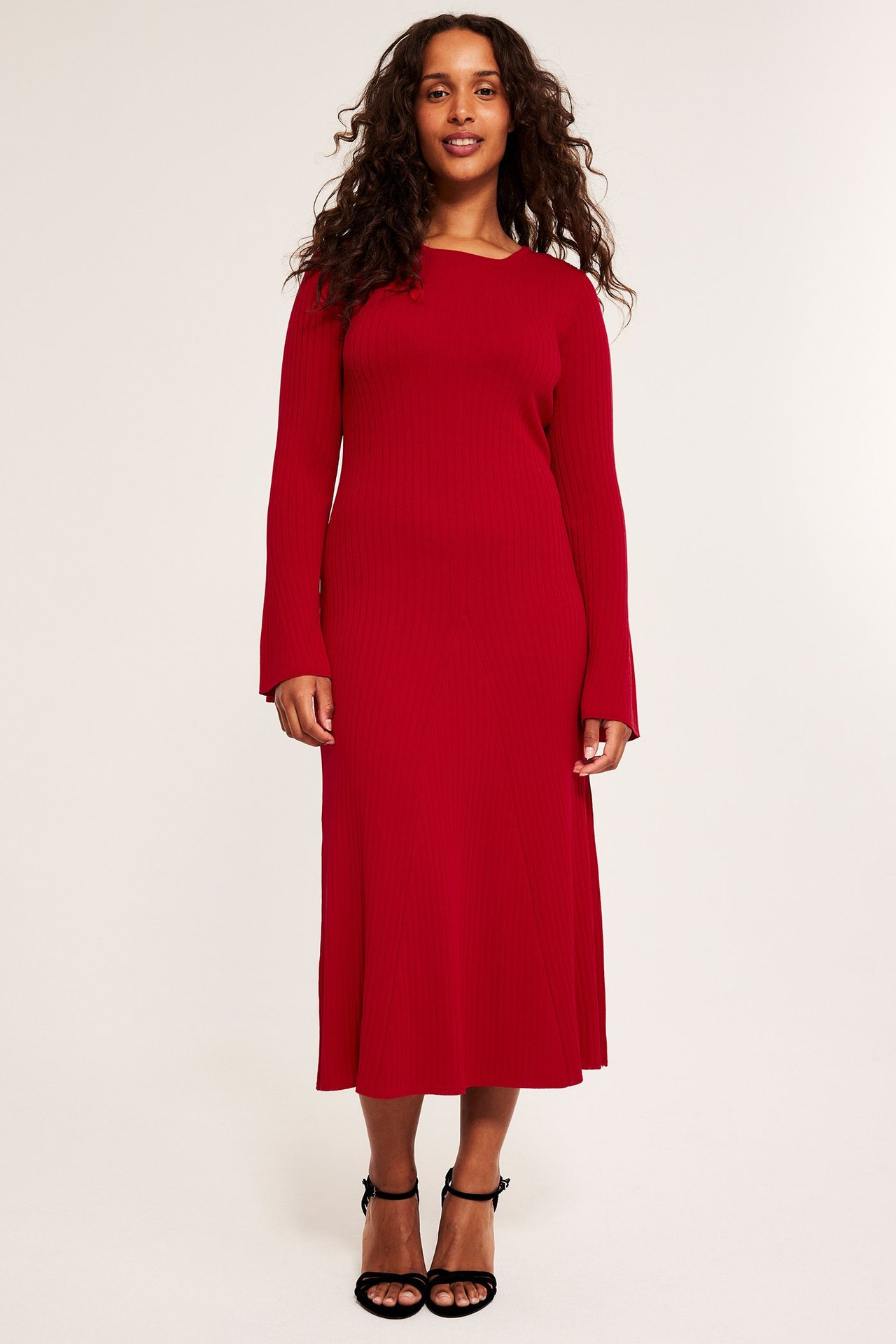 Frontvy av en kvinna i röd stickad midiklänning med långa utställda ärmar och svarta högklackade sandaler.