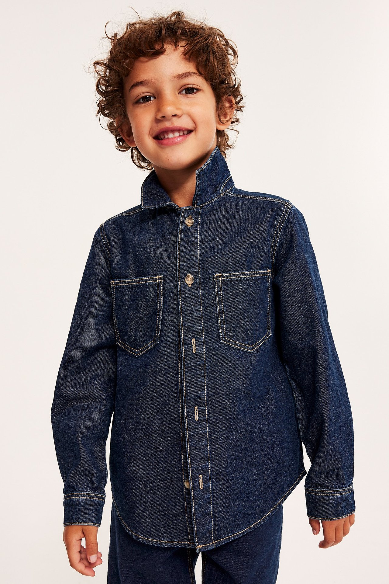 Barn stående framifrån med kort lockigt brunt hår. Klädd i stängd mörkblå jacka i denim med knappar framtill och bröstfickor på vardera sida. Matchande mörka jeans nedtill. Pojken ler.