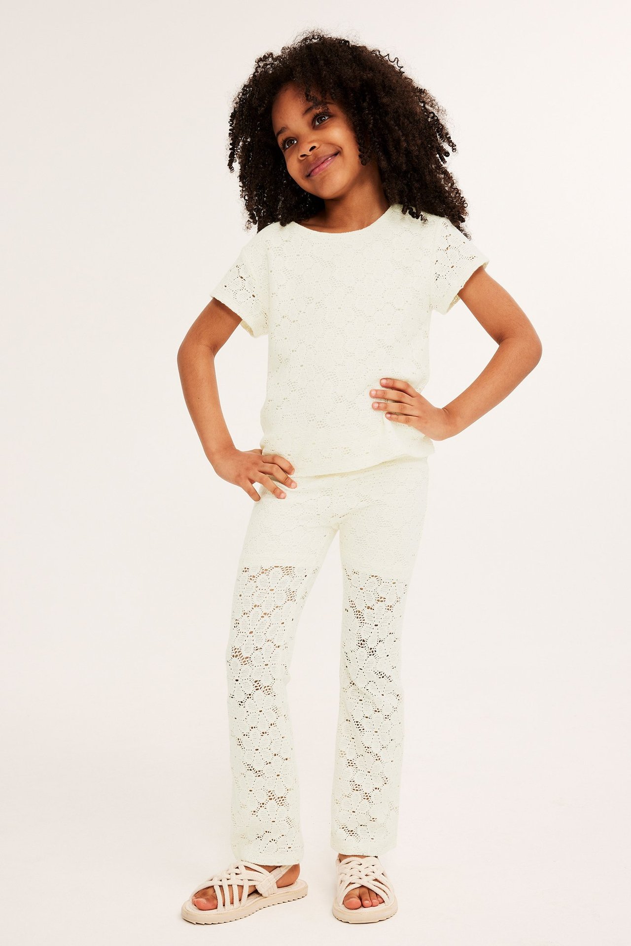Dziecko w kremowym koronkowym topie, pasujących spodniach i jasnych sandałach.