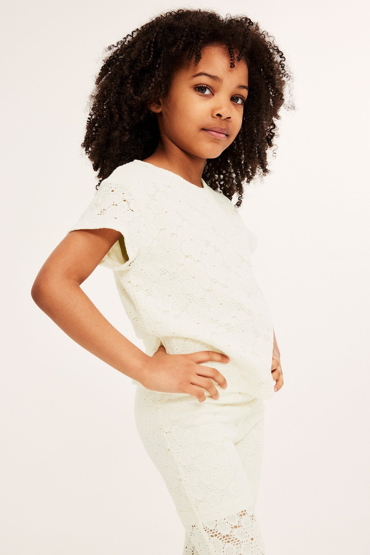 Widok z przodu dziecka w kremowym koronkowym topie z krótkim rękawem i pasujących koronkowych spodniach.