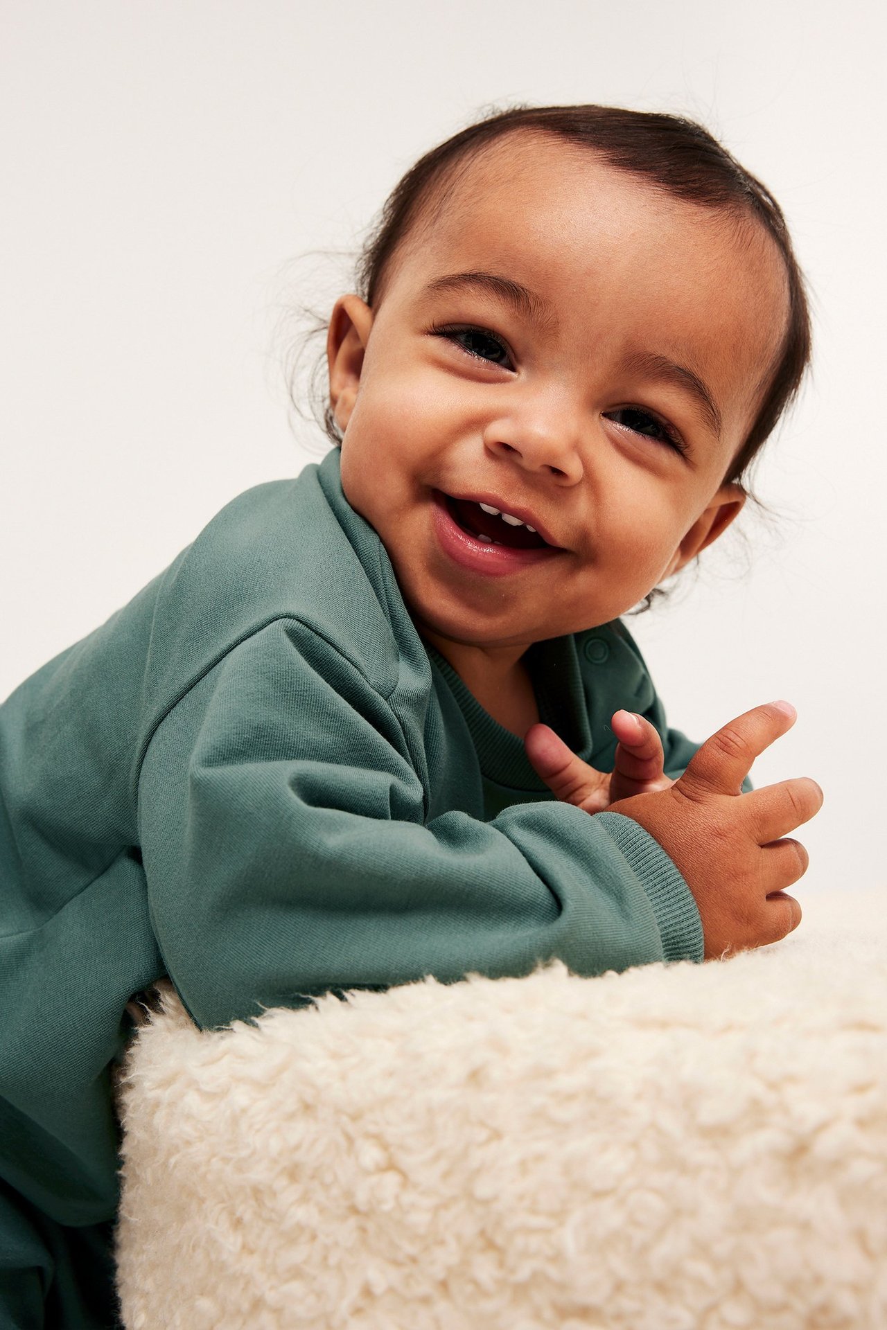 Edestä kuvattu hymyilevä vauva yllään khakinvihreä puuvillainen collegepaita resoreilla.