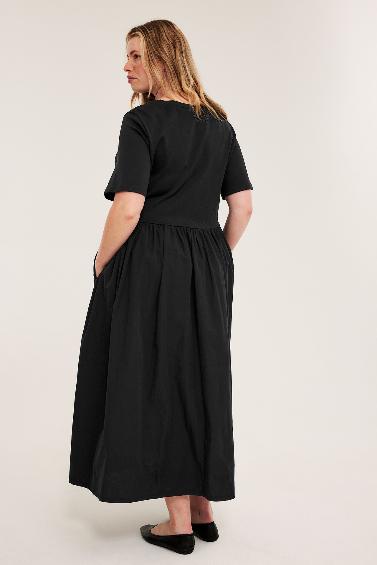 Kvinna, bakifrån, i en svart kortärmad lång klänning med rynkad midja och svarta platta skor.