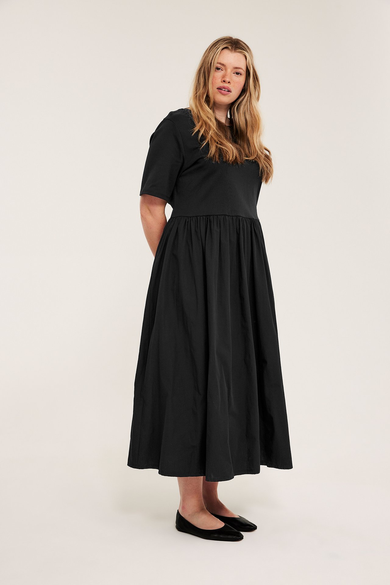 Front view: Kvinna i svart rundhalsad kortärmad maxiklänning, poplinkjol, och svarta ballerinaskor.