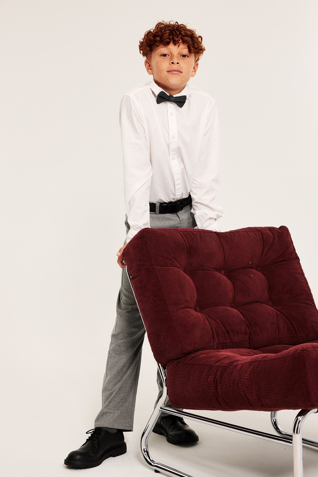 Widok z przodu dziecka w białej koszuli, czarnej muszce, szarych spodniach, czarnym pasku i butach.