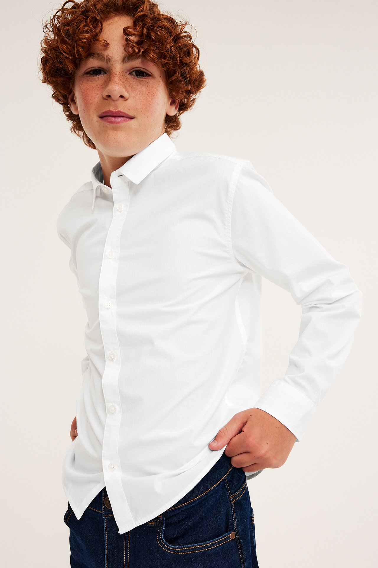 Edestäpäin kuvattu lapsi valkoisessa slim fit -pitkähihaisessa paidassa ja tummansinisissä farkuissa.