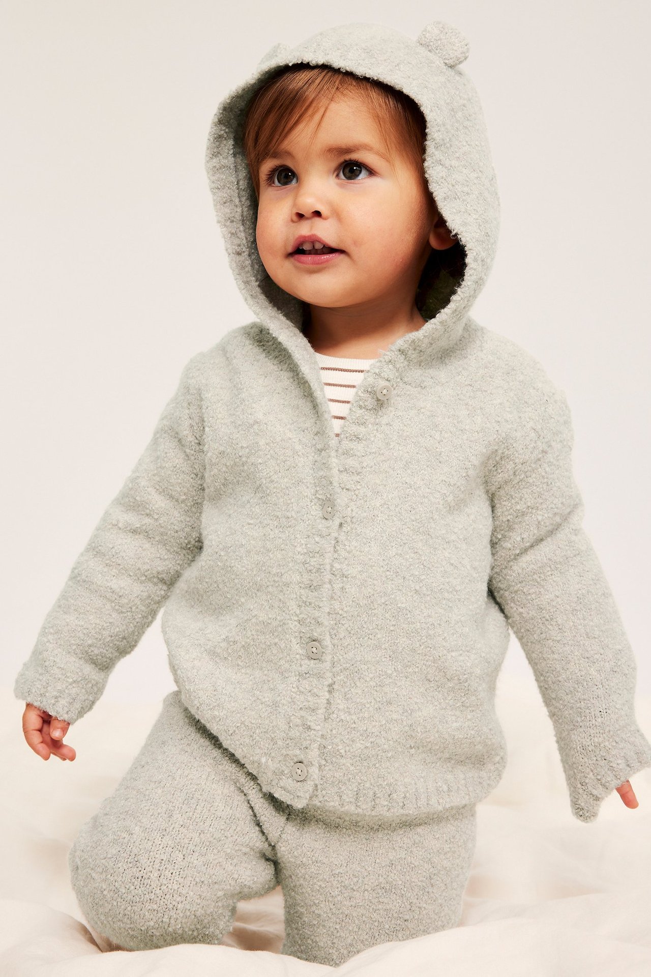 Framifrån, en baby i en beigemelange boucléstickad kofta med huva med björnöron och matchande byxor.
