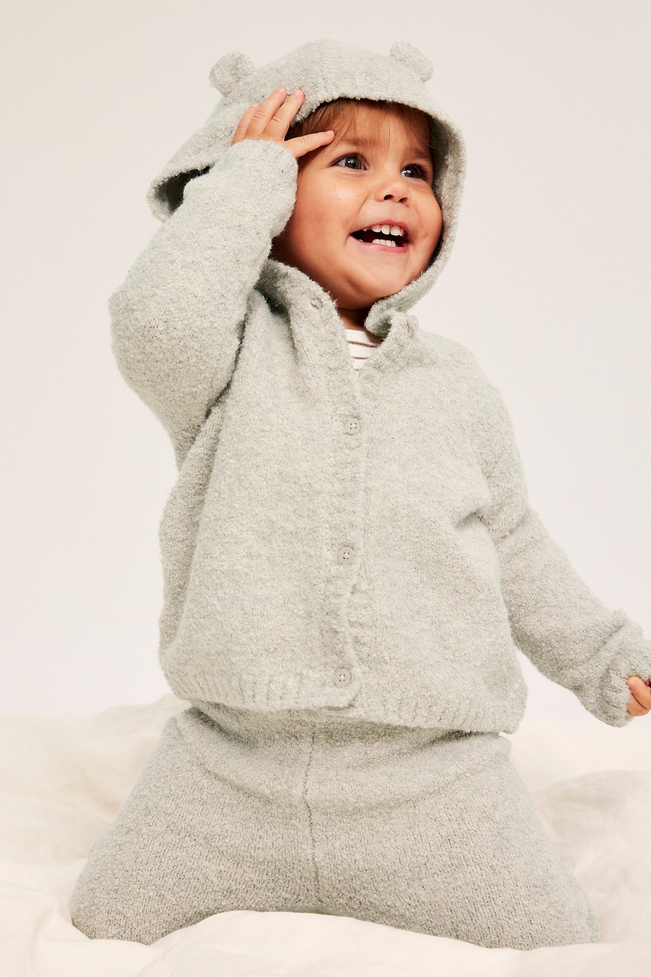 Widok z przodu: Uśmiechnięte dziecko w beżowym melanżowym kardiganie buclé z kapturem, uszkami i pasującymi spodniami.