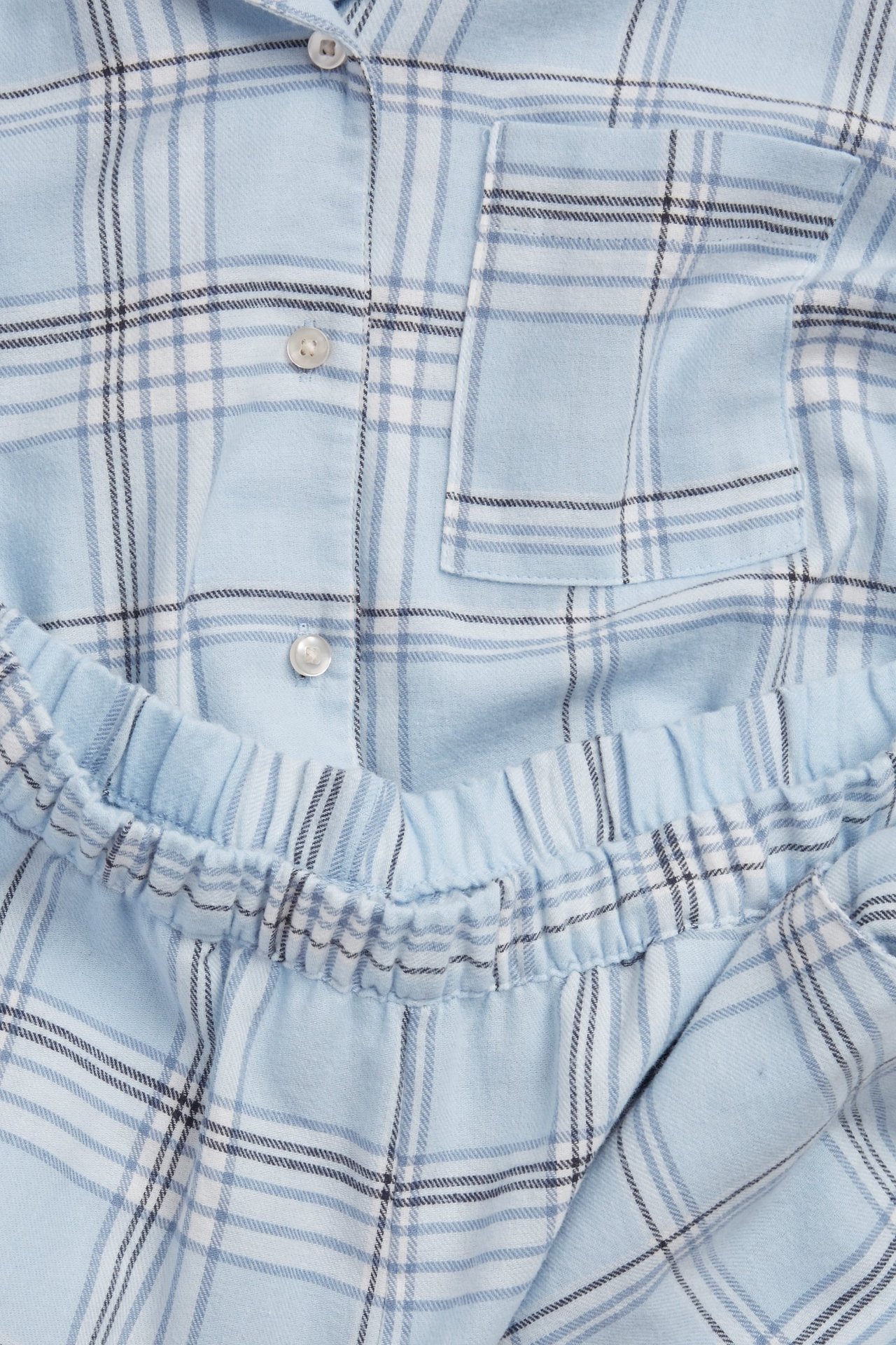 Nærbilde av lyseblå rutete flanellpyjamas med knapper, brystlomme og elastisk midje.