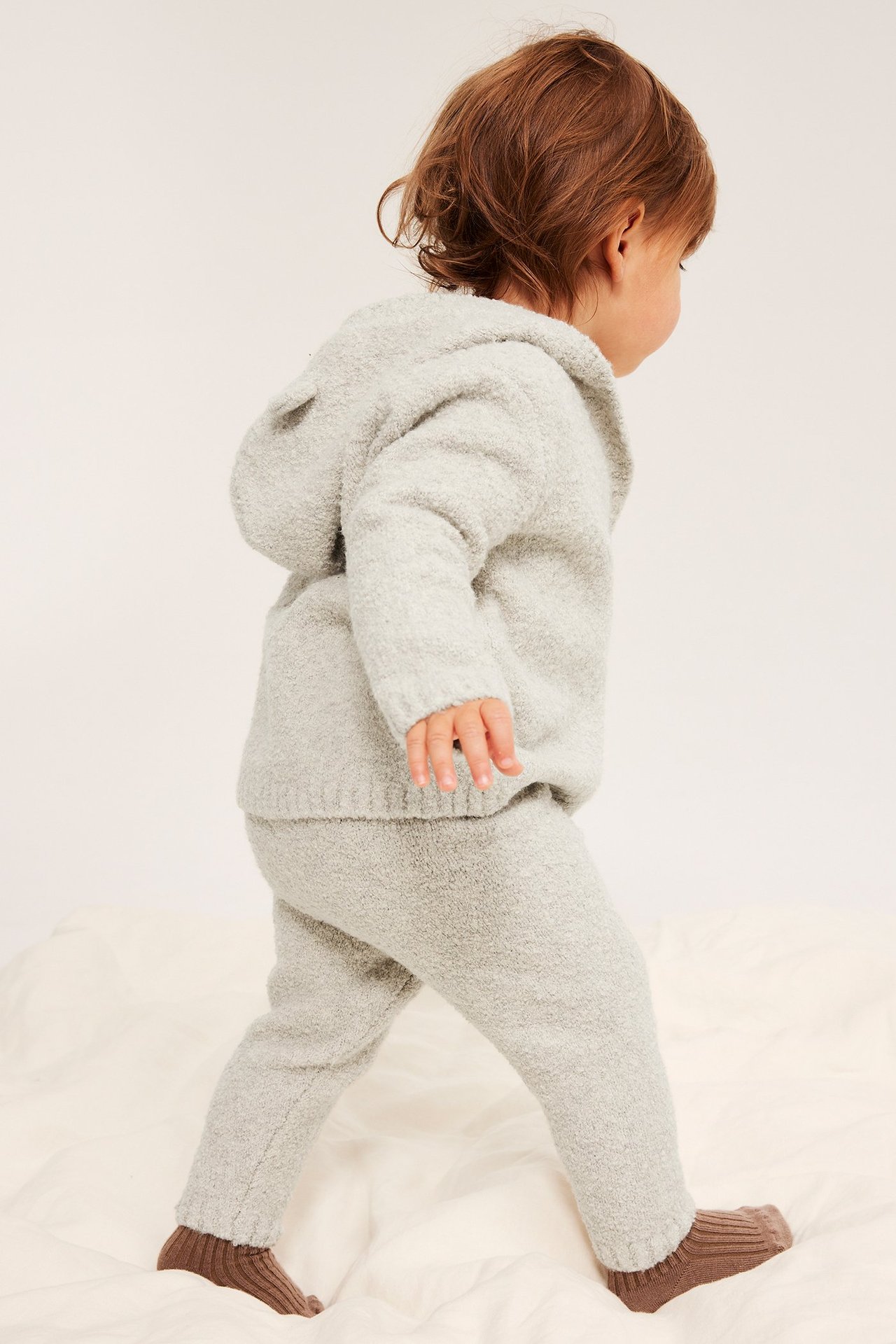 Sned bakifrån-vy av baby i beigemelange boucléstickade leggings, matchande huvtröja med öron och bruna strumpor.